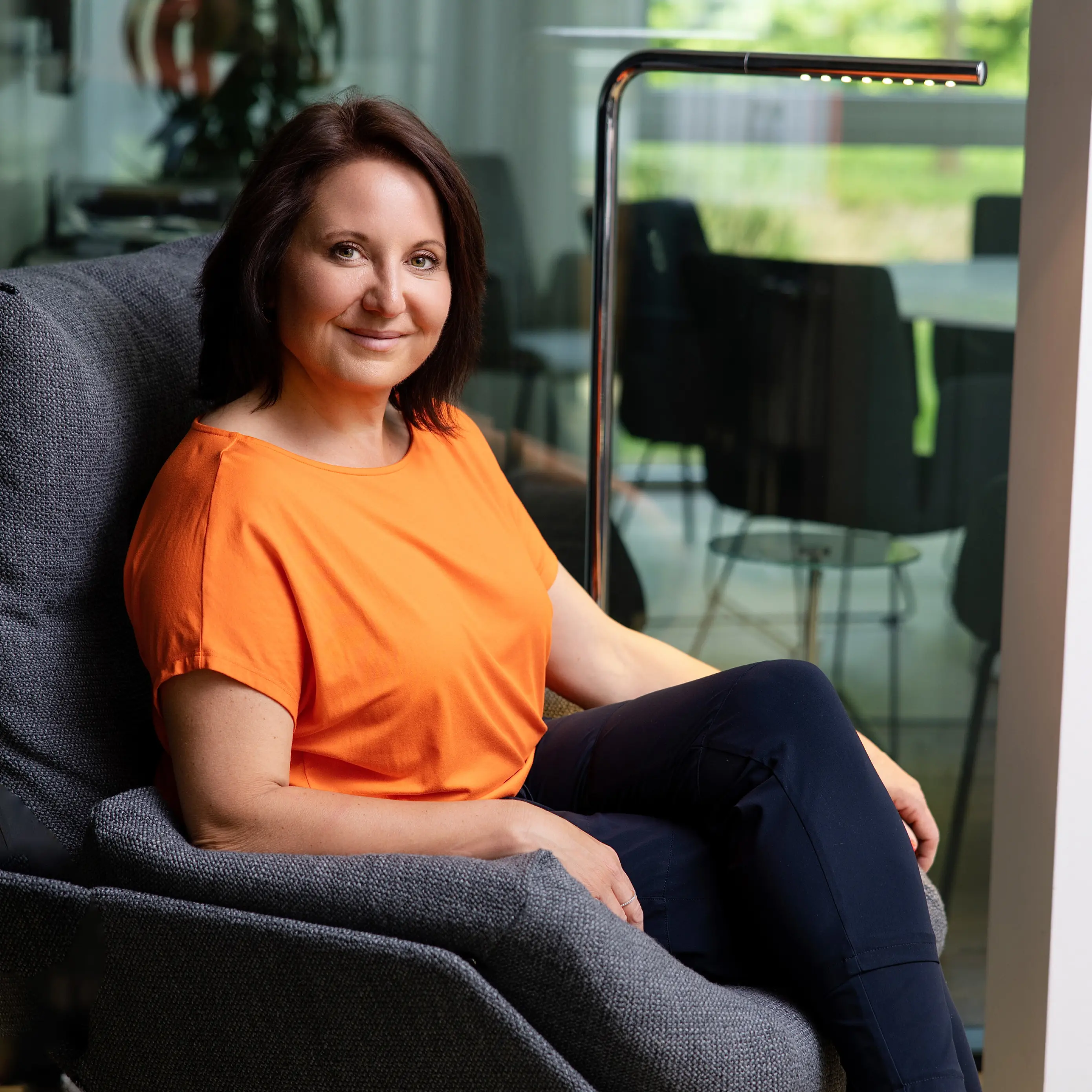 Marion Mossmann Schwarz sitzt in orangefarbenem Oberteil in einem Sessel und blickt in die Kamera.