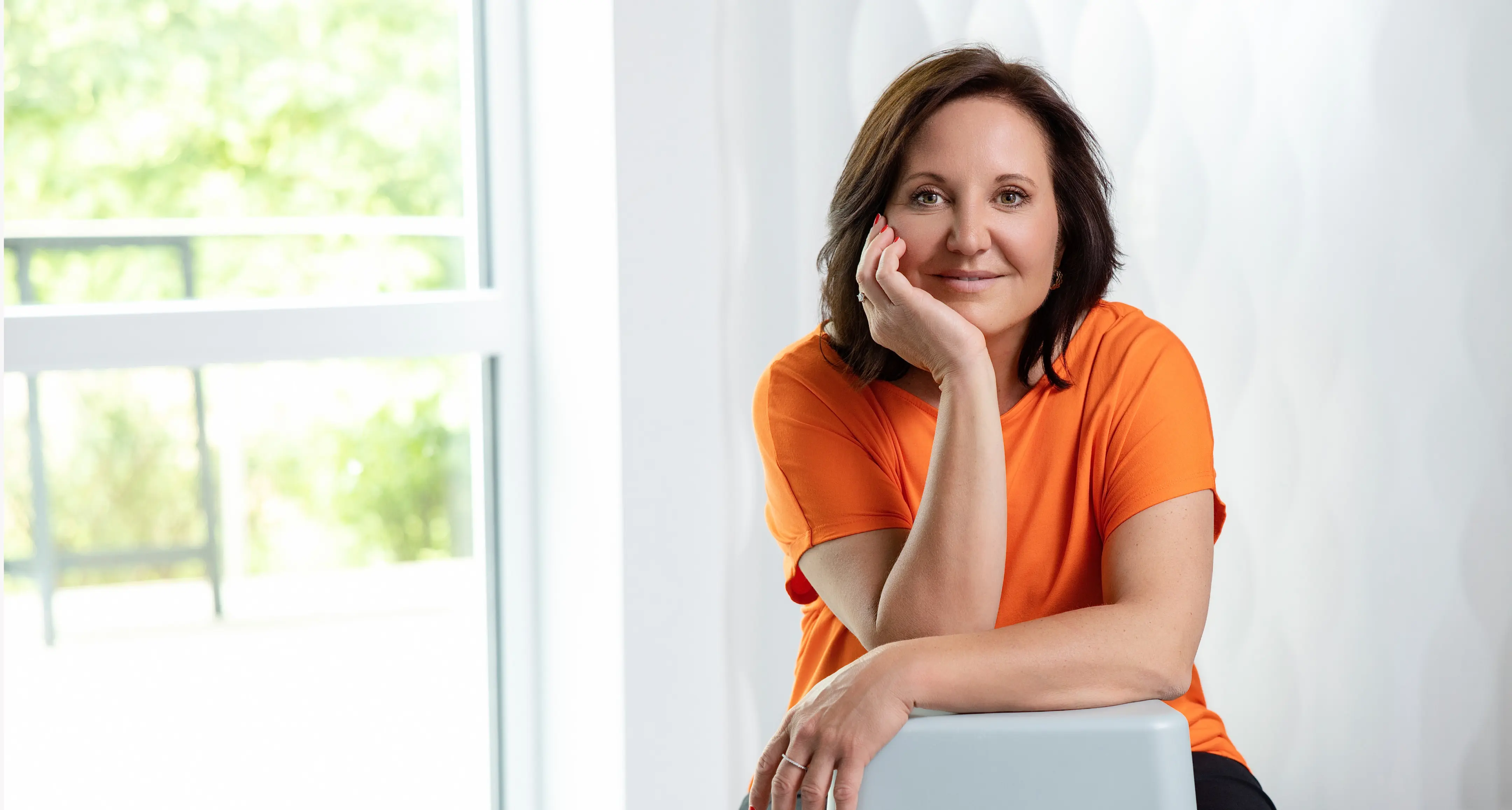 Marion Mossmann Schwarz lehnt in orangefarbenem Oberteil an einem hellen Arbeitsplatz und blickt in die Kamera.