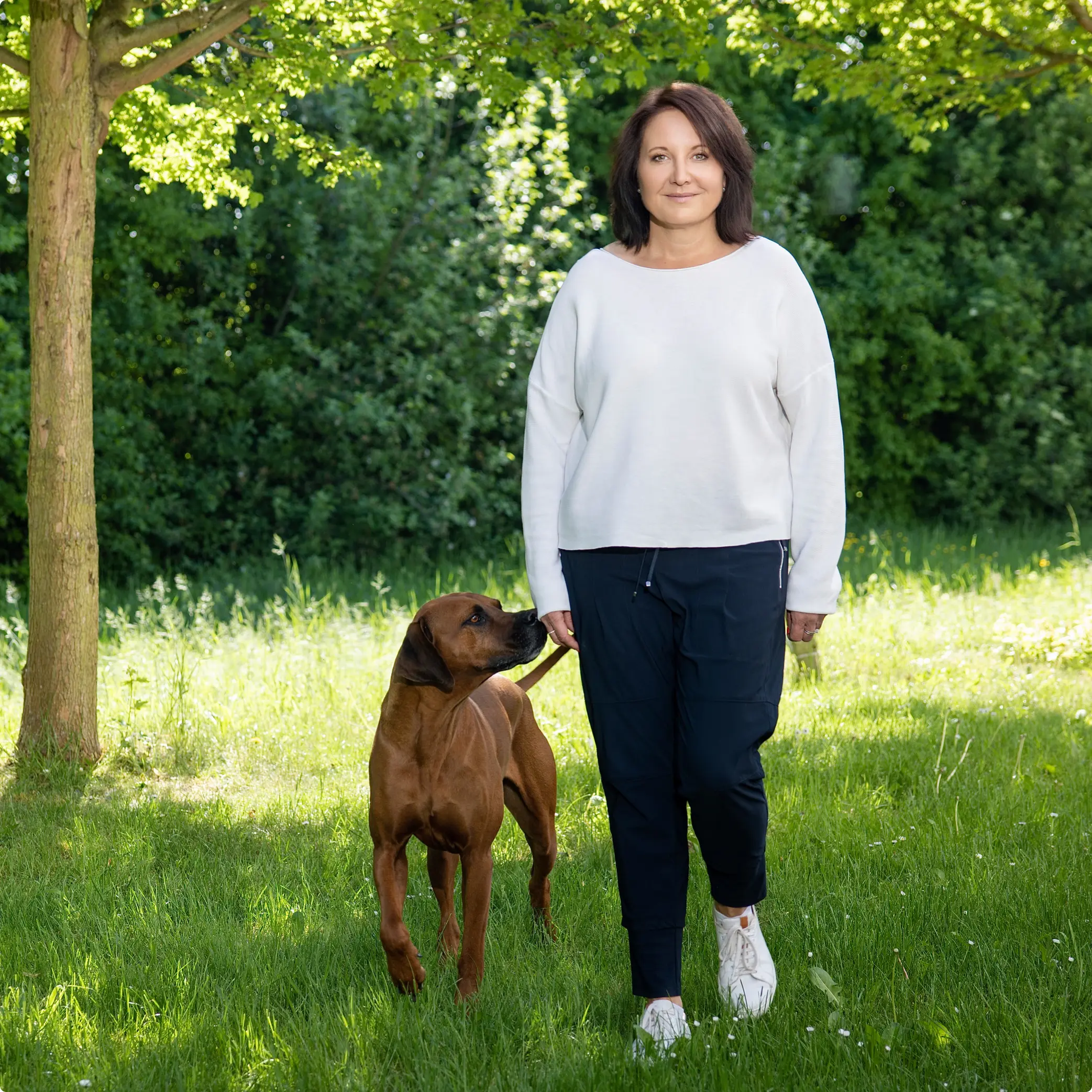 Marion Mossmann Schwarz steht mit einem Hund auf einer grünen Wiese unter Bäumen.