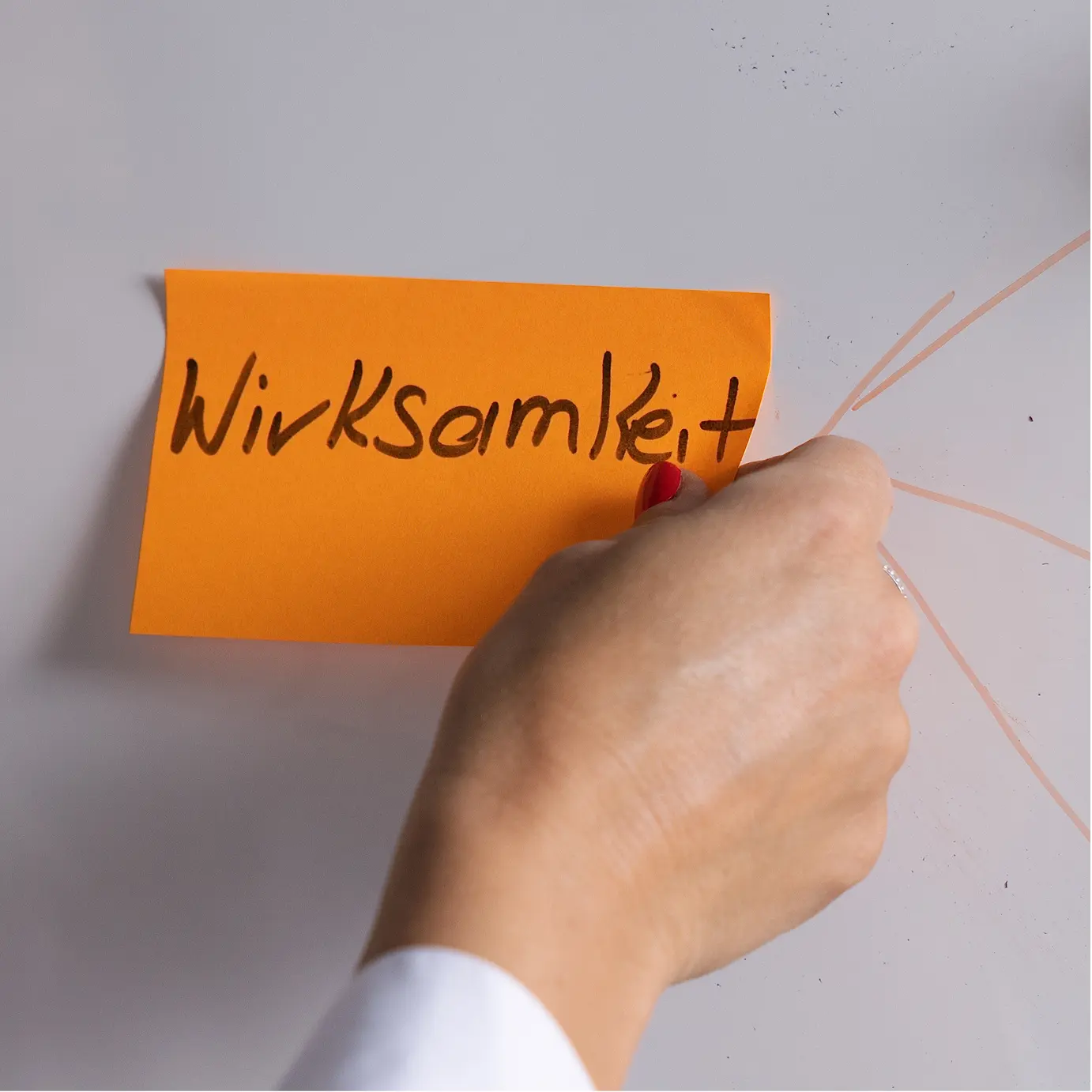 Hand schreibt auf ein orangefarbenes Post-it mit dem Wort Wirksamkeit als Symbol für messbaren Nutzen von Wissen.