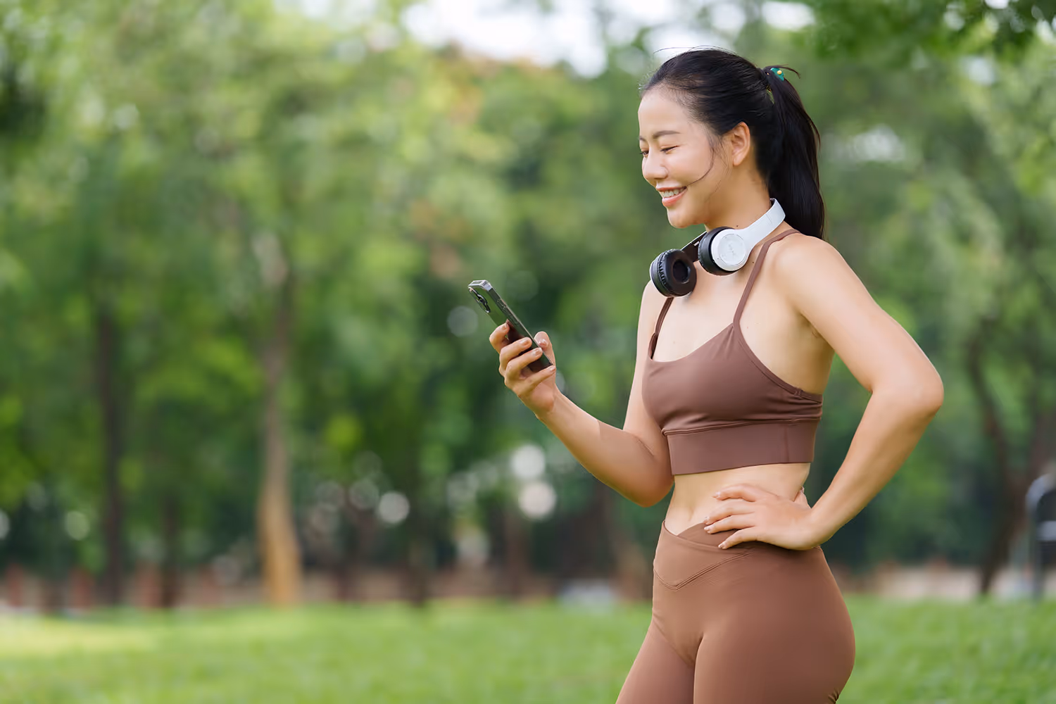 Frau in Sportkleidung mit Kopfhörern schaut lächelnd auf ihr Handy im Park.