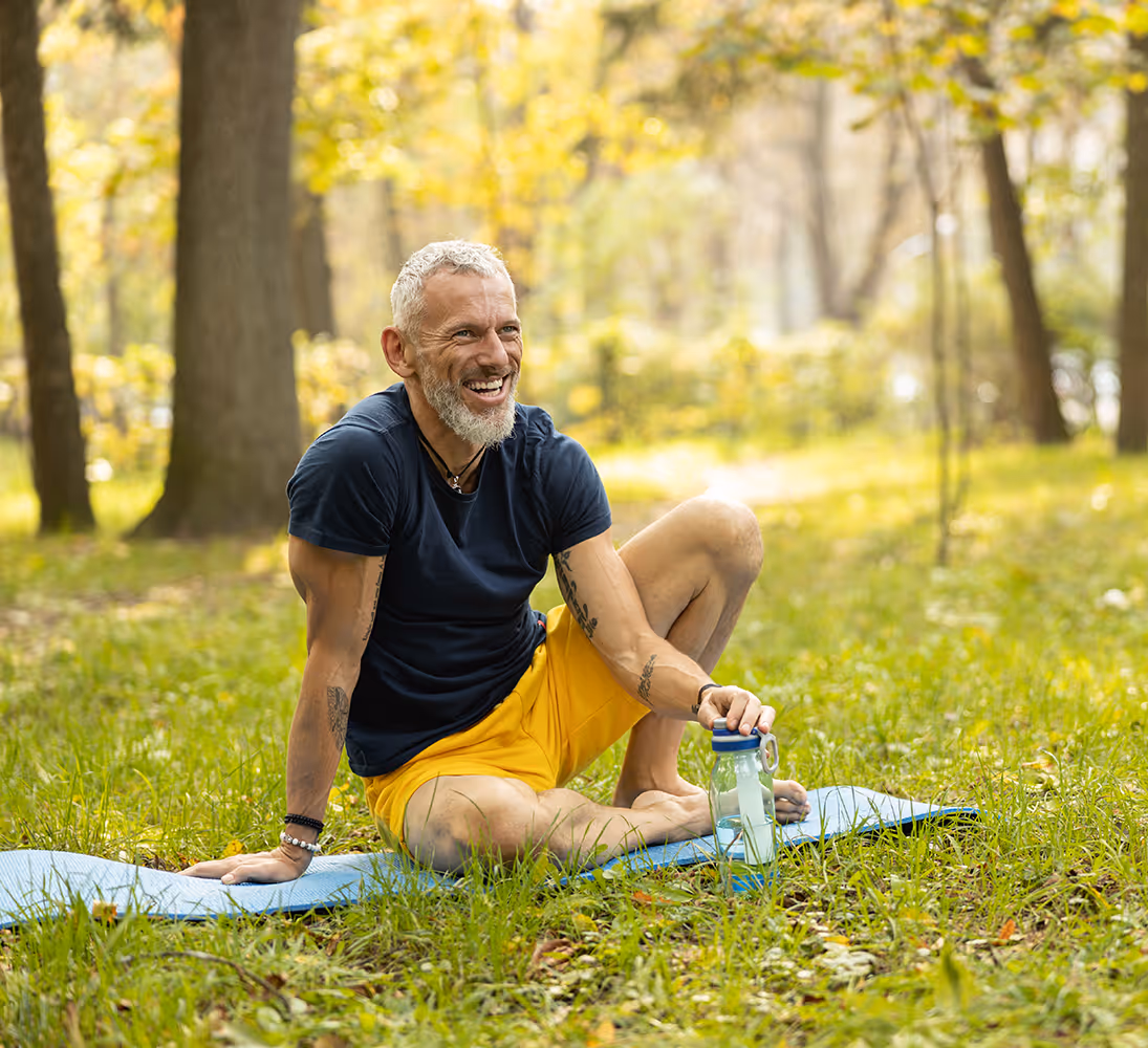Älterer Mann mit Bart in gelben Shorts sitzt auf Yogamatte im Park und lächelt, hält Trinkflasche.