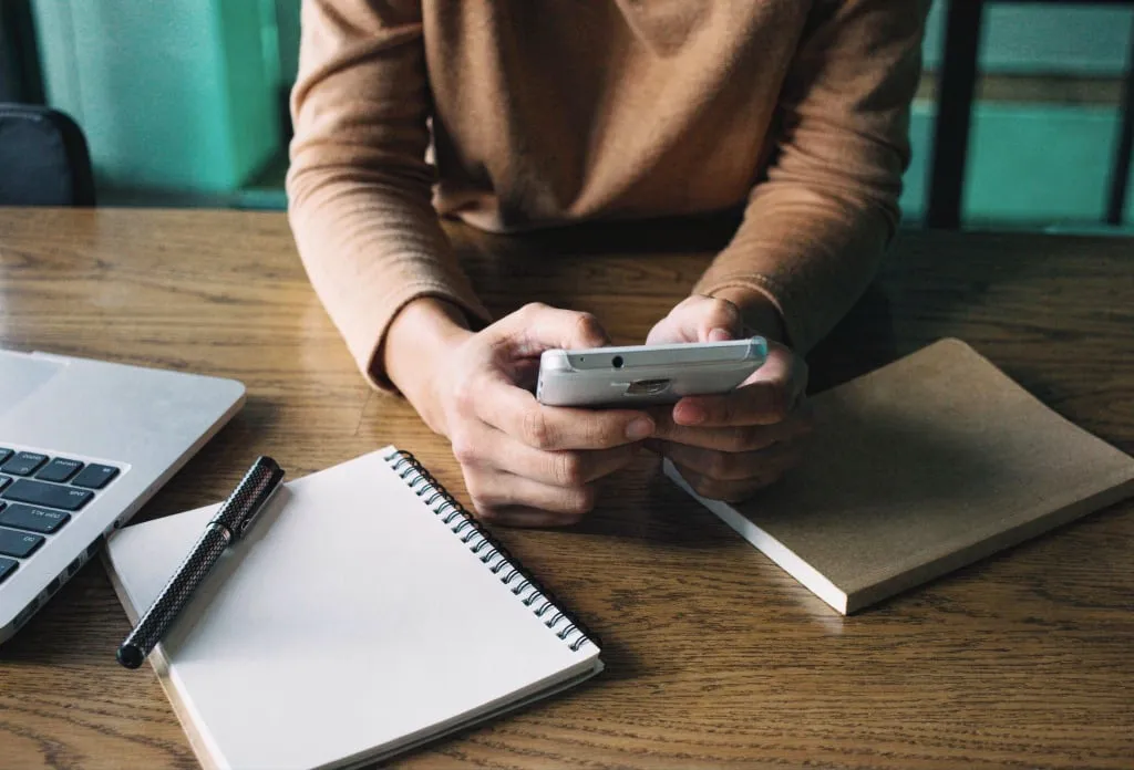 Persona con suéter marrón usando un teléfono móvil mientras están sentados en una mesa de madera con un cuaderno blanco de espiral, un bolígrafo, una libreta marrón y una computadora portátil parcial visible.