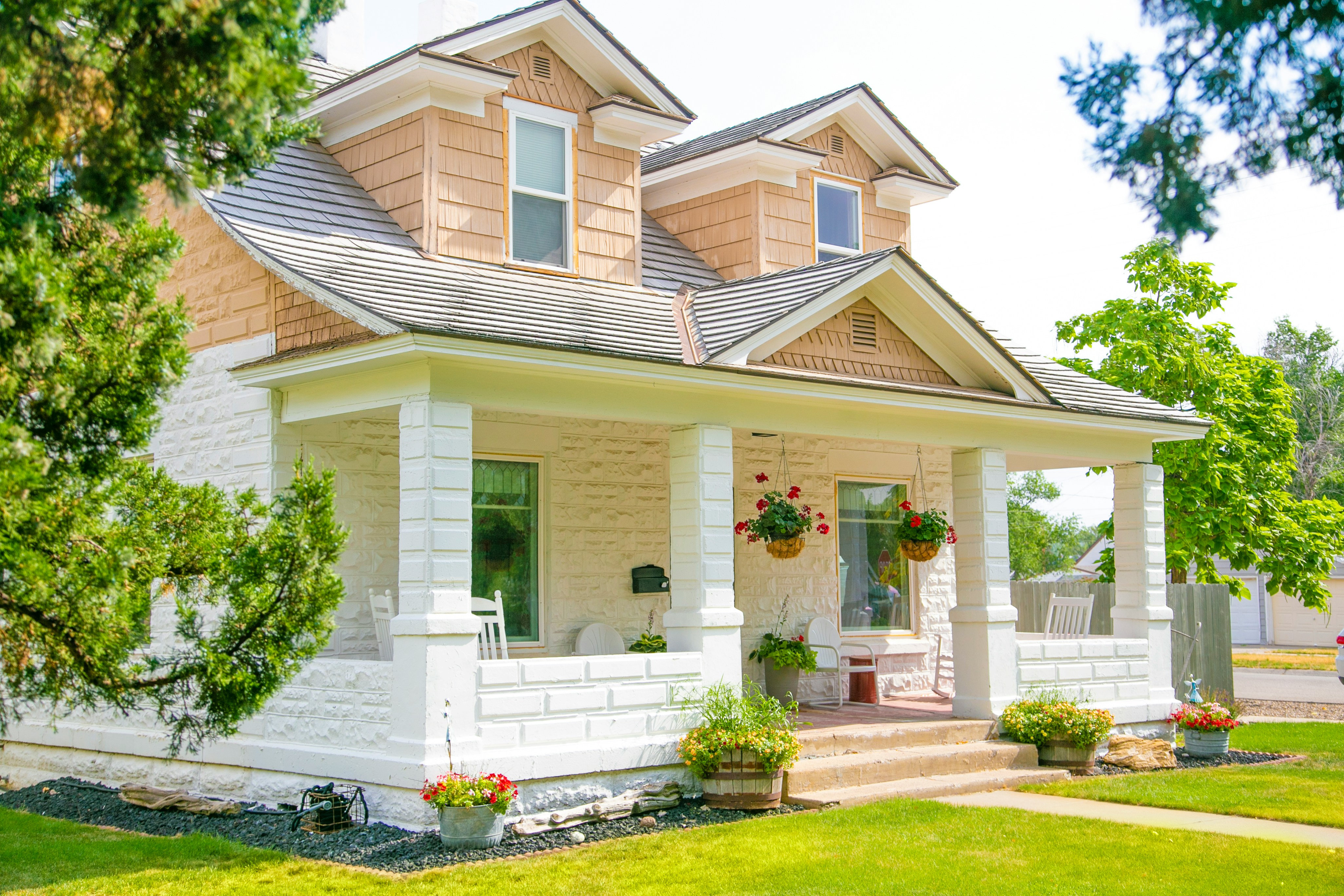 A white and tan craftsman-style house with a covered front porch, white columns, hanging flower baskets and a green front lawn.
