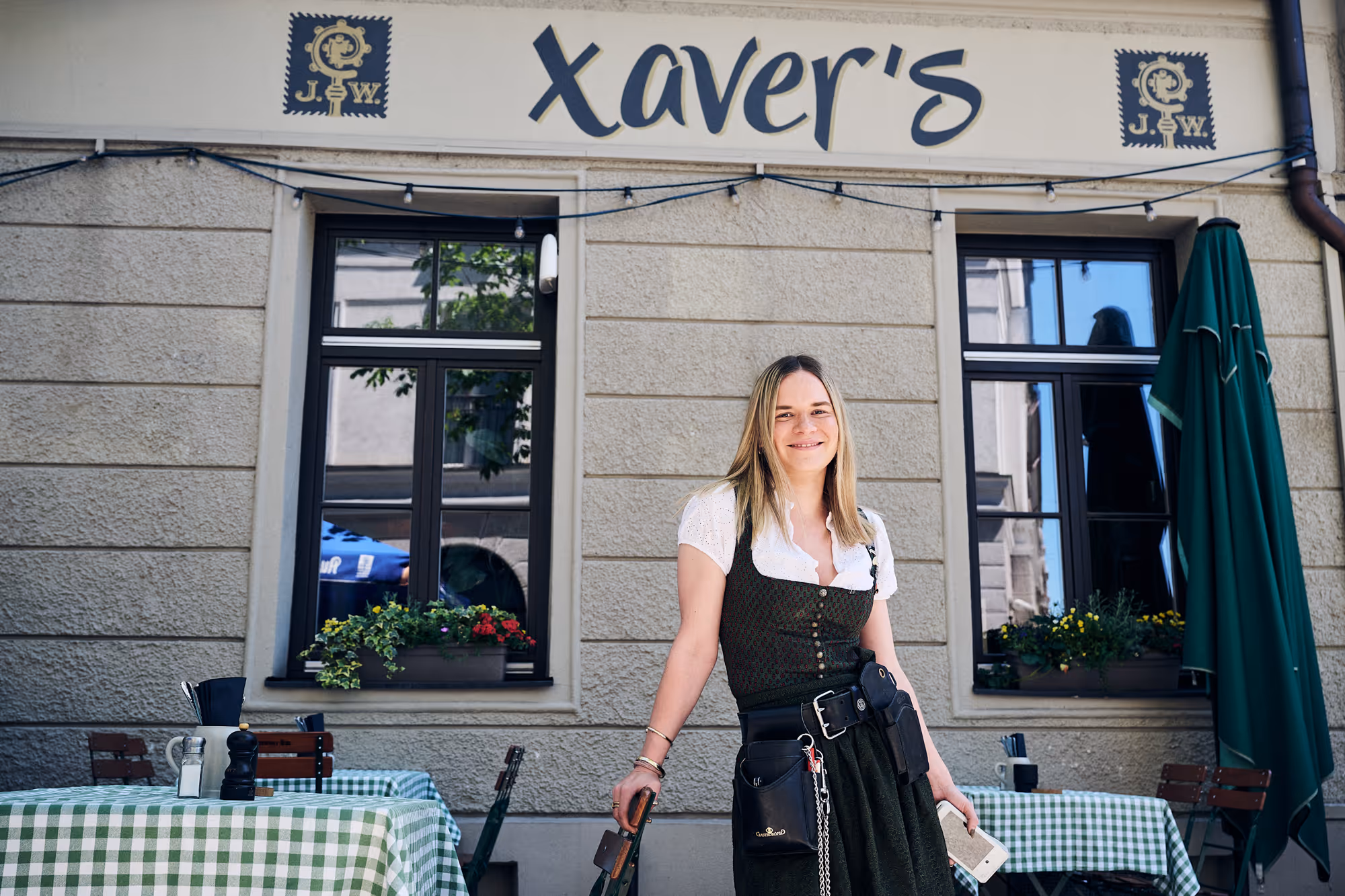 Servicekraft in Tracht vor der Fassade des Restaurants Xaver's mit Außenbestuhlung