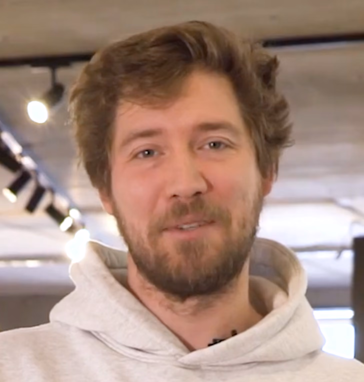 Ein junger Mann mit braunen Haaren und Bart trägt einen hellen Kapuzenpullover und lächelt.