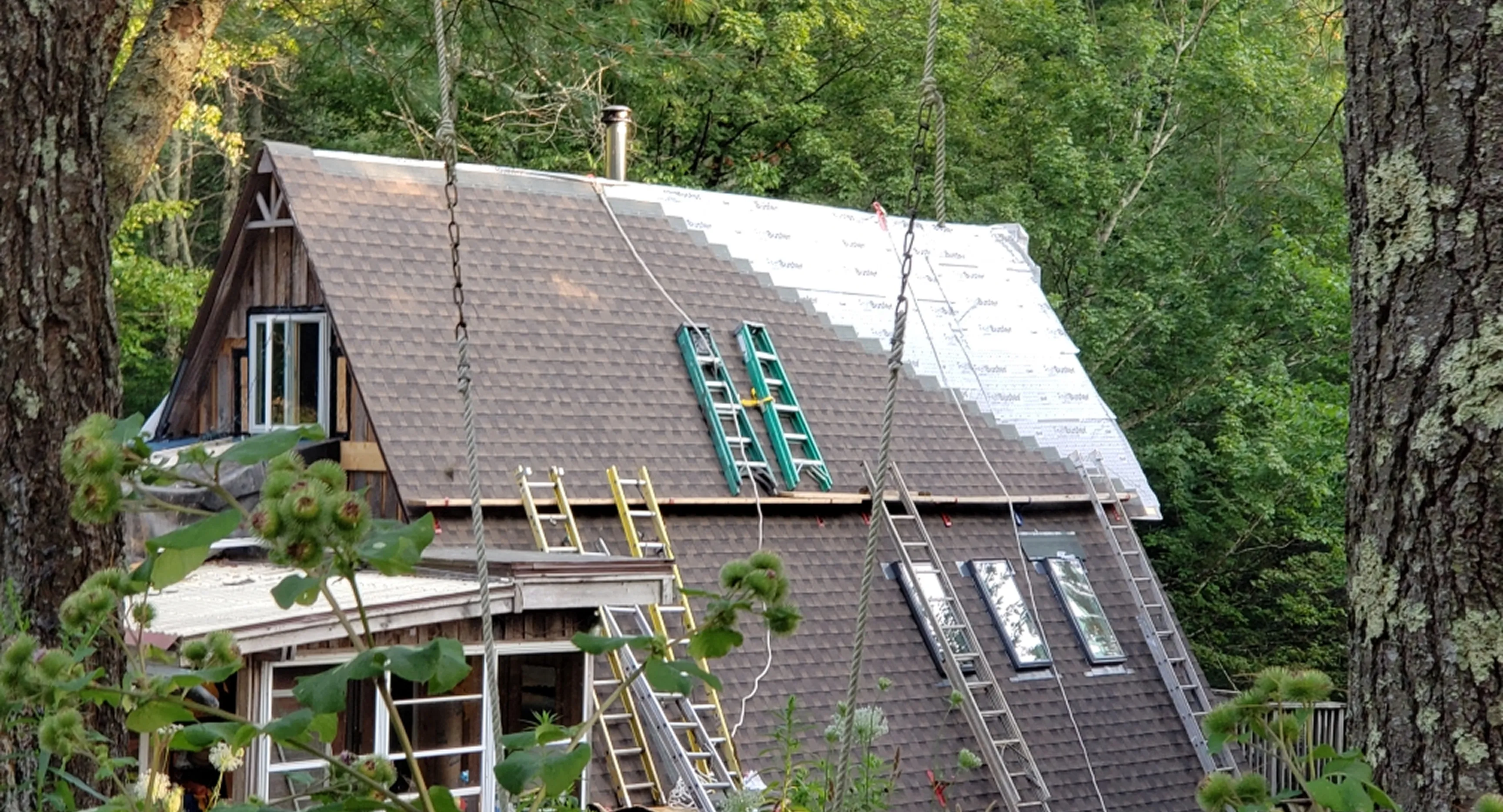 A-frame house roof under construction with ladders and shingles, surrounded by trees and plants.