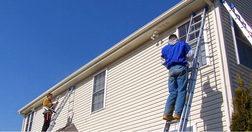 Two workers climbing ladders against a beige house under a clear blue sky.