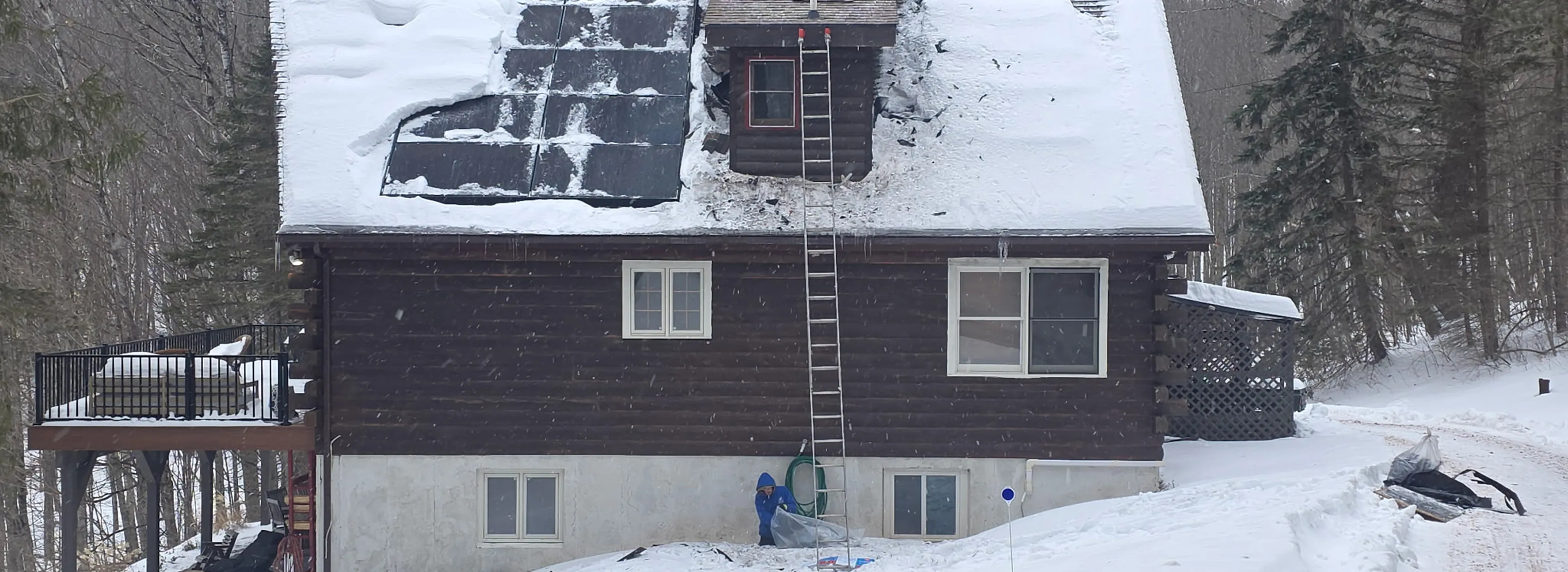 Snow-covered dark wooden house with solar panels on roof and a person working near the foundation.