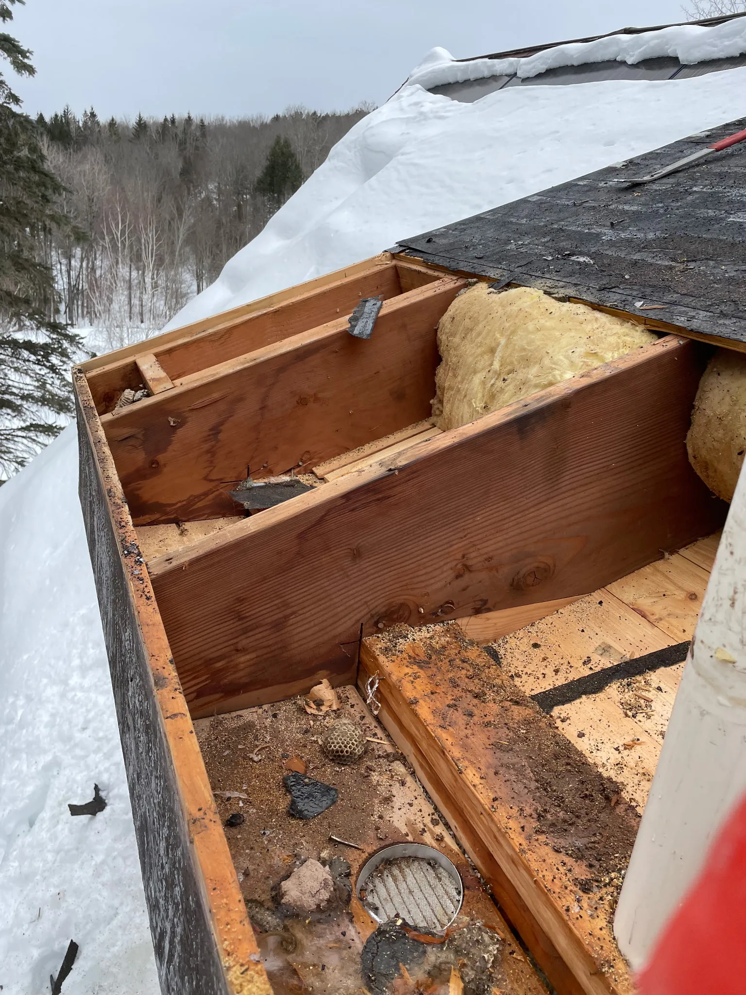 Partially removed roof section showing wooden beams, insulation, debris, and snow on roof edge.