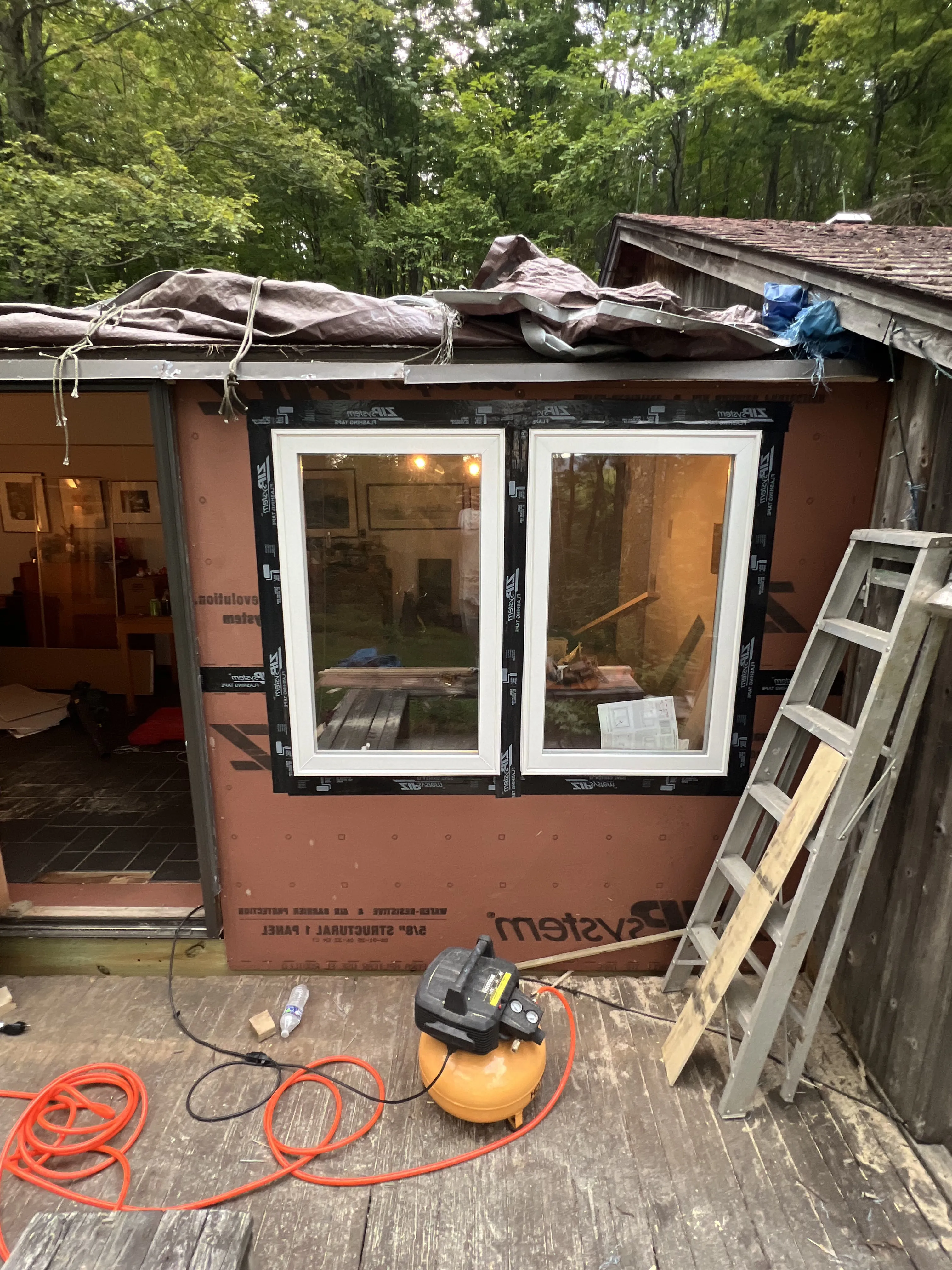 Construction area with two newly installed white-framed windows, ladder, air compressor, and tools on wooden deck.