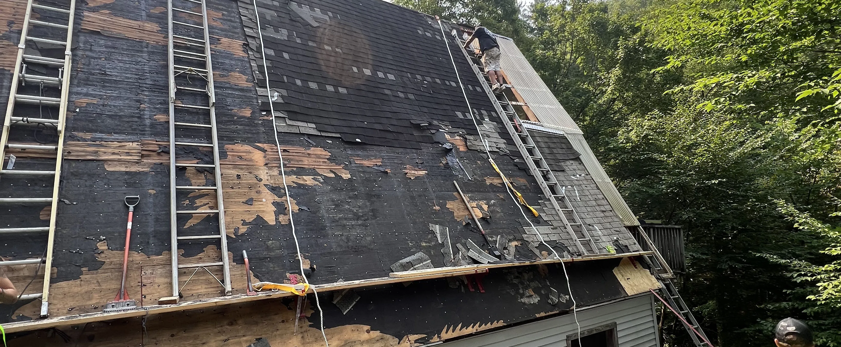 Construction worker on ladder repairing a roof with missing shingles near a wooded area.