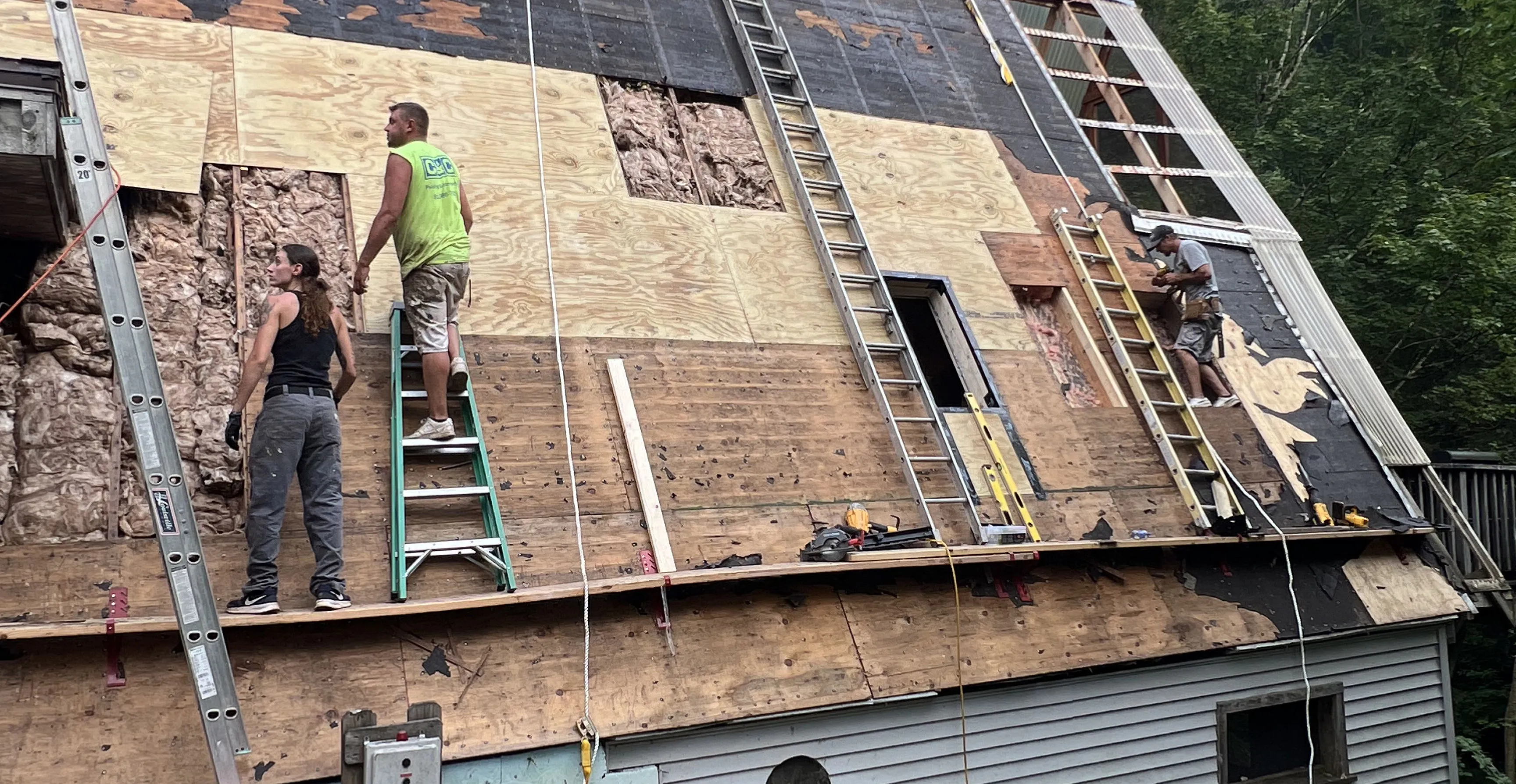 Three workers on ladders installing plywood and insulation on a steep roof under construction.