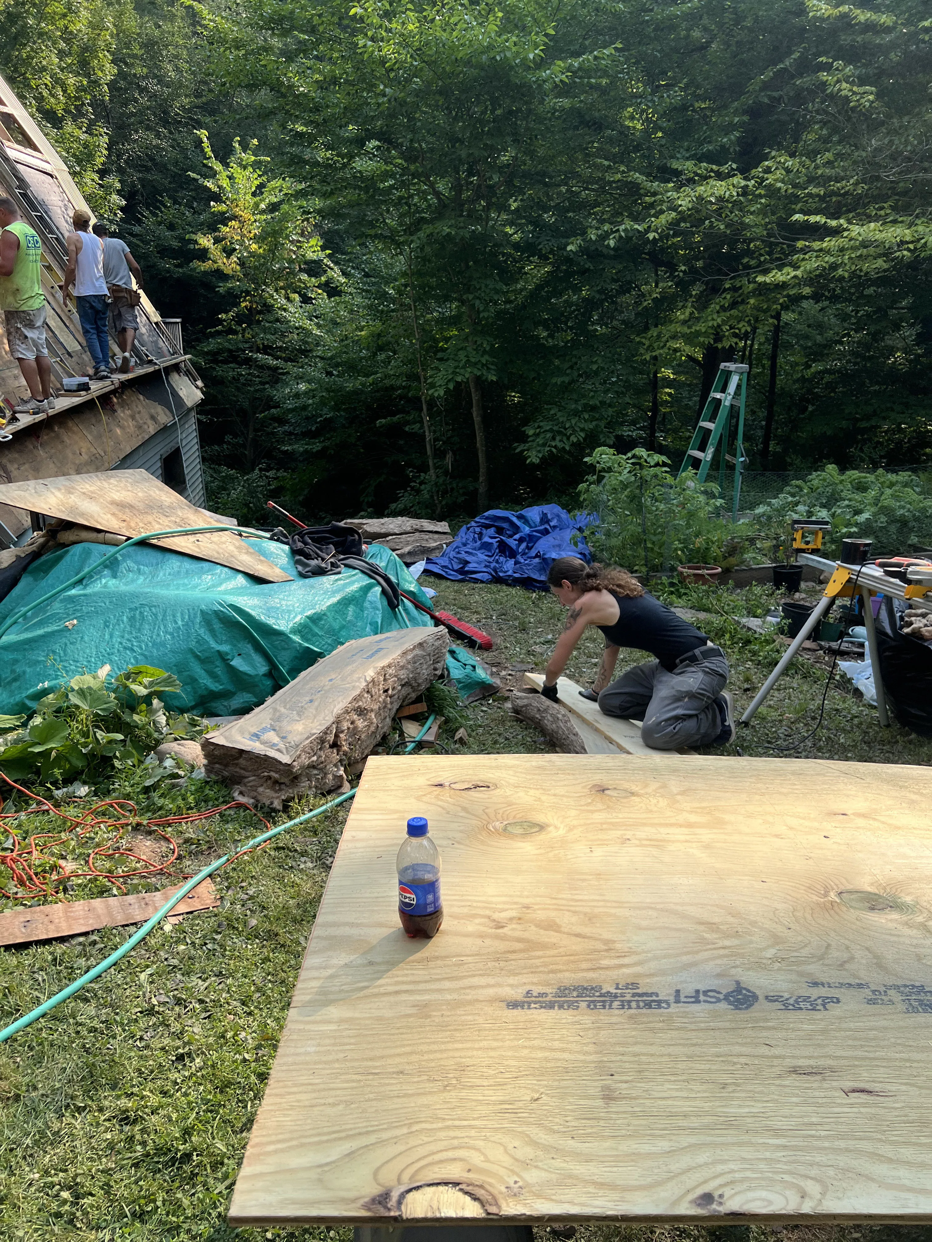 Person kneeling near wooden boards outdoors with three workers on a roof and construction tools nearby.