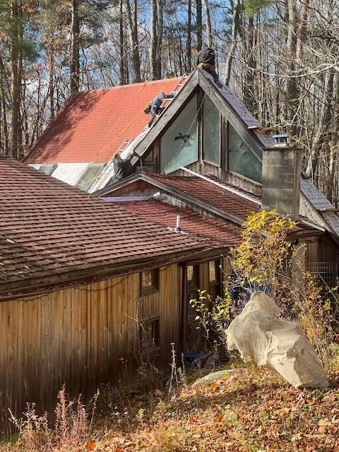 Workers installing red roofing shingles on a rustic cabin surrounded by autumn trees.