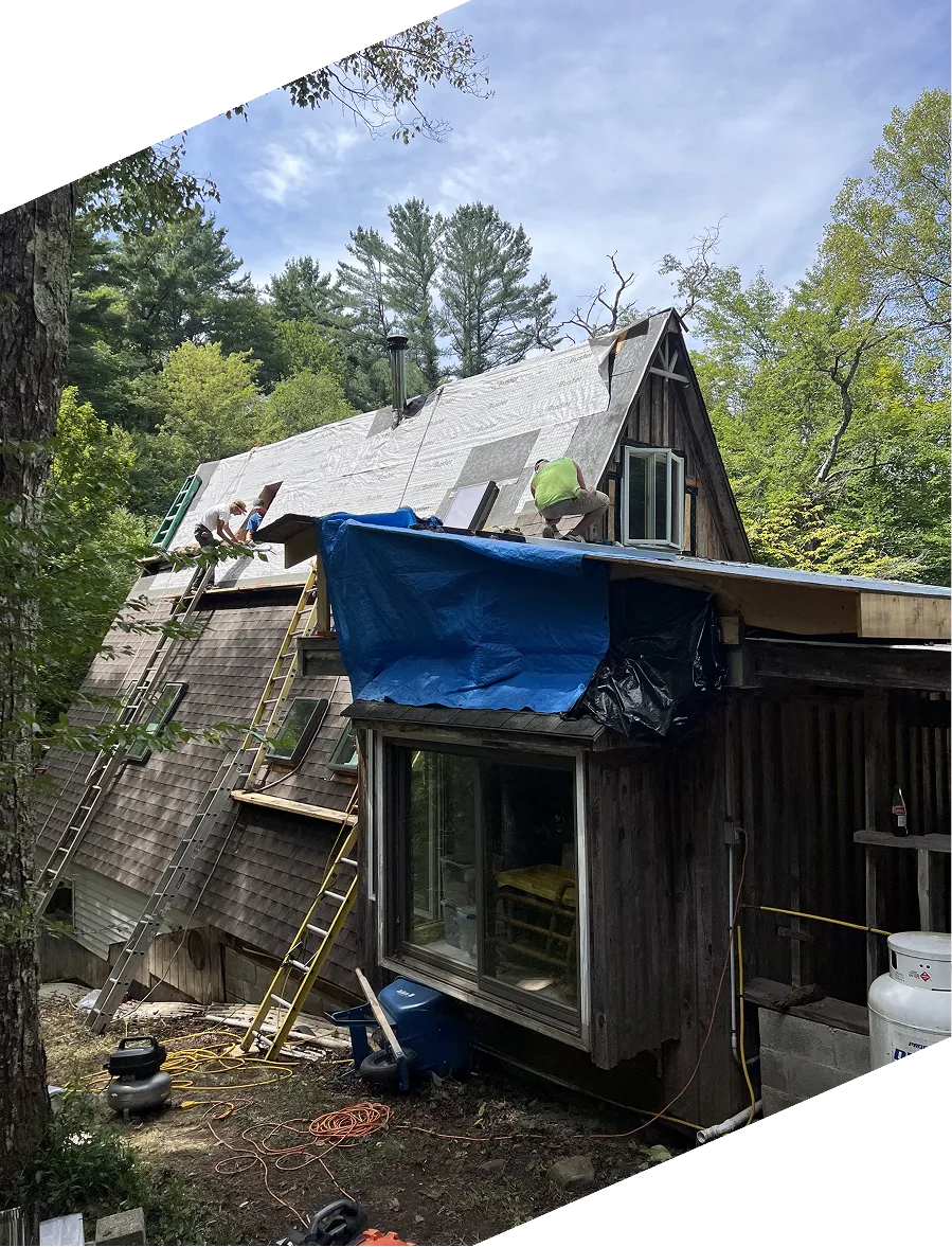 Workers on ladders installing roofing underlayment on an A-frame cabin surrounded by trees.
