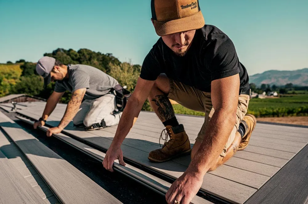 Two men installing wooden deck boards outdoors with tools and boots under clear sky.