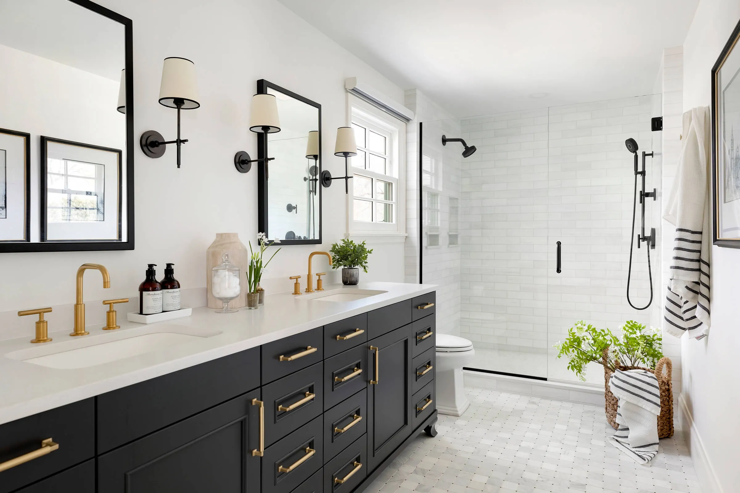 Modern bathroom with black cabinets, gold fixtures, dual sinks, walk-in shower, and white tiled flooring.
