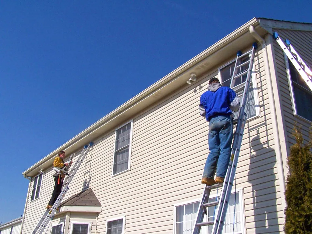 Two workers on ladders installing or repairing beige siding on a two-story house under clear blue sky.