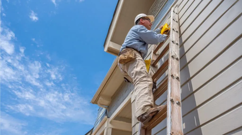 Worker in protective gear climbing a wooden ladder and inspecting house siding under blue sky.