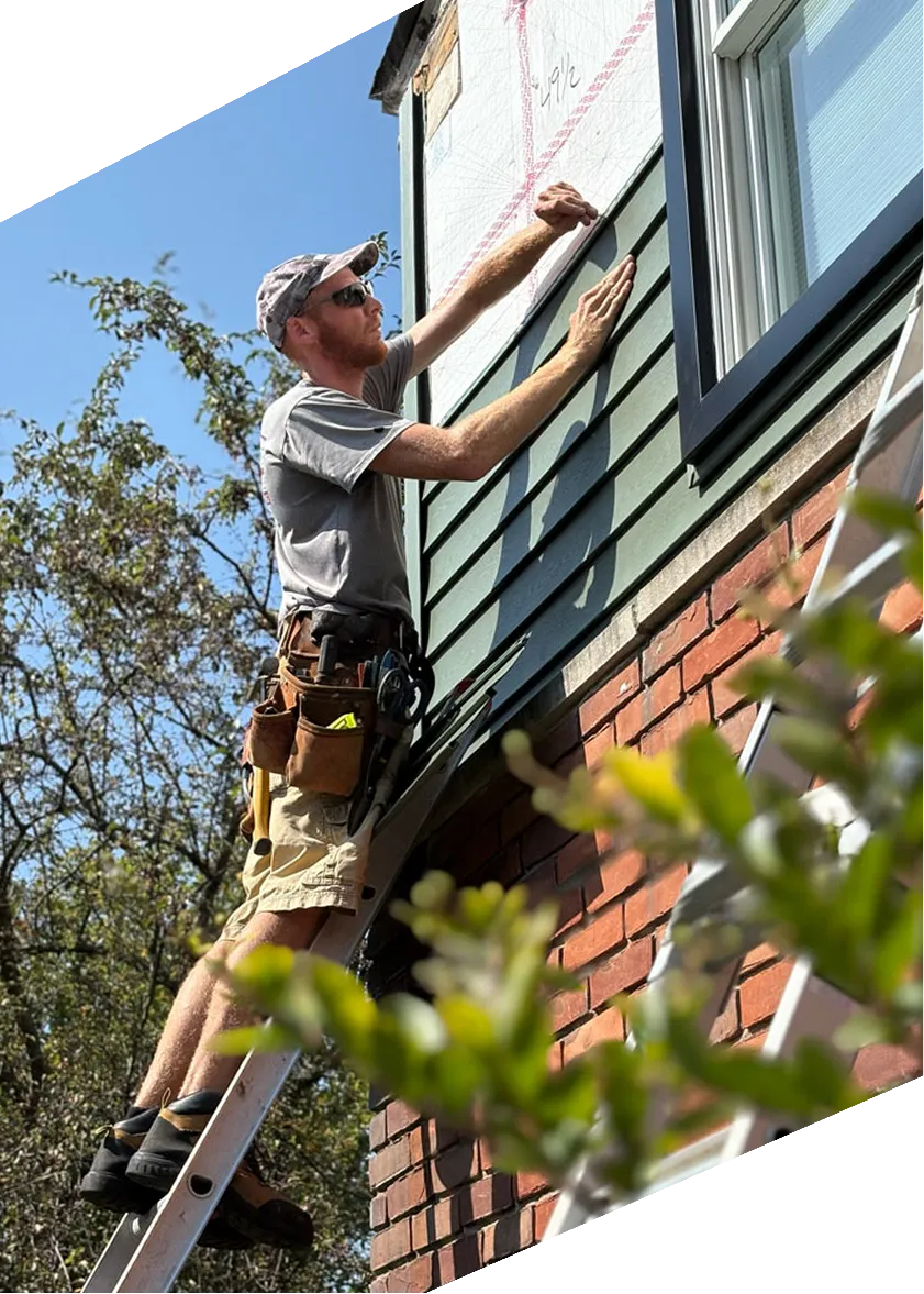 Construction worker on ladder installing green siding on a brick house under a clear blue sky.