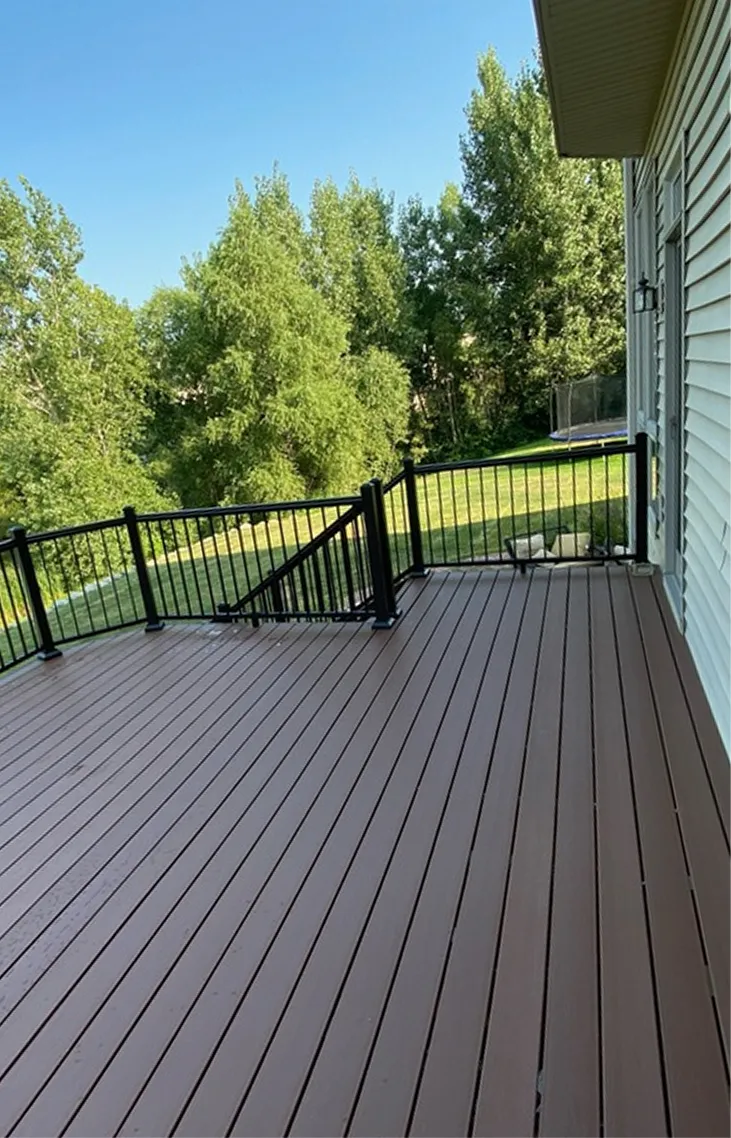 Spacious wooden deck with black railing attached to a white house overlooking green yard and trees.