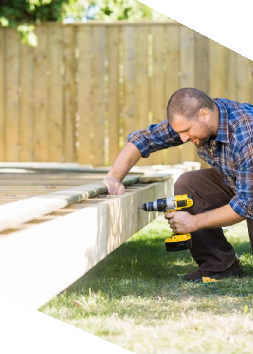 Man using a cordless drill to build a wooden deck outdoors in a grassy yard.