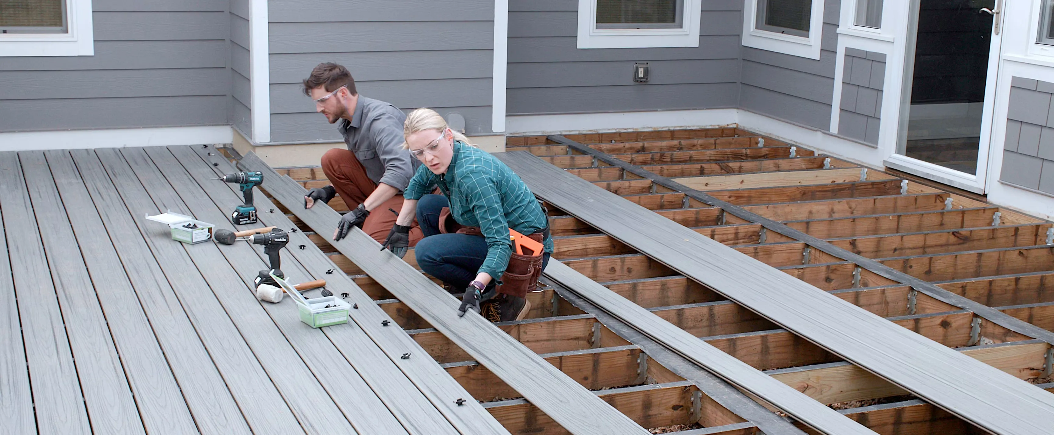 Two people wearing safety glasses installing gray wooden deck boards on a wooden frame outside a house.