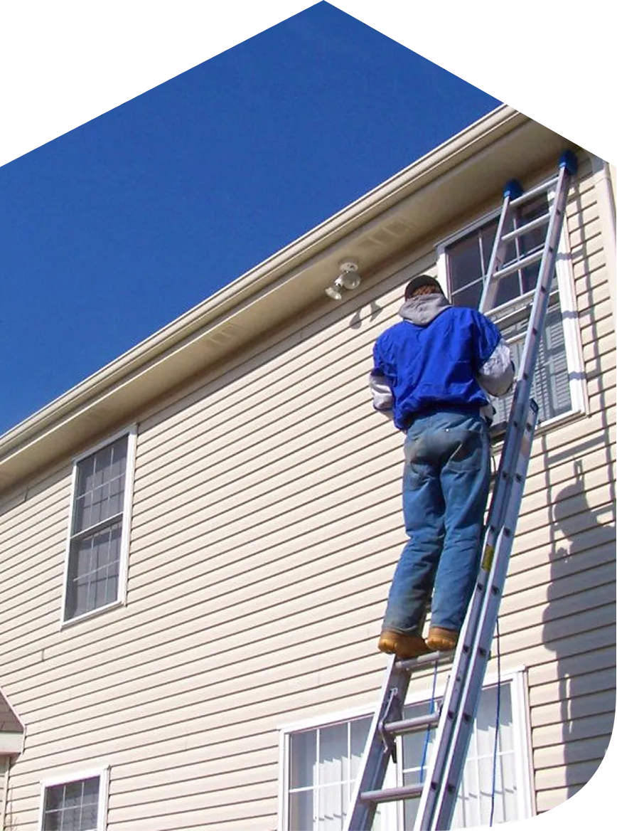 Worker in blue jacket climbing ladder to install or repair beige vinyl siding on house under clear blue sky.