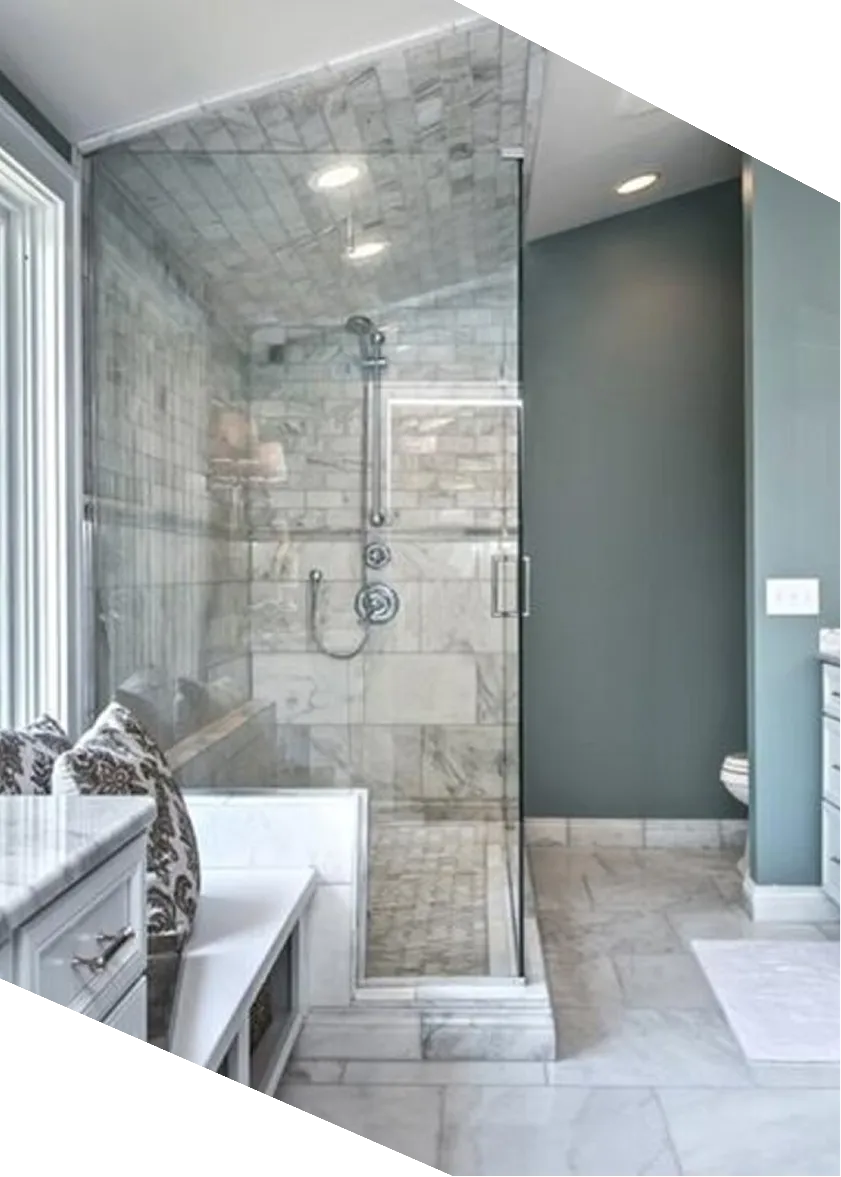 Modern bathroom with glass shower, marble tiles, bench with patterned pillows, and gray-green walls.