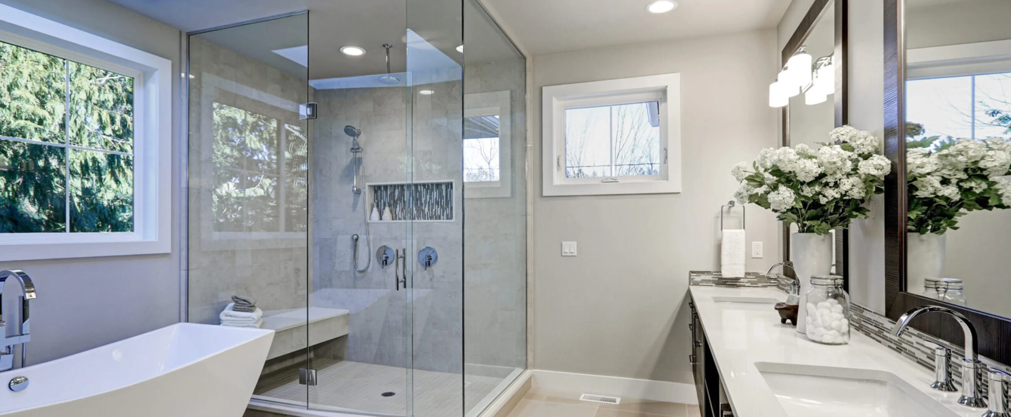 Modern bathroom with glass shower, freestanding tub, double sink, and white floral arrangement by the mirror.
