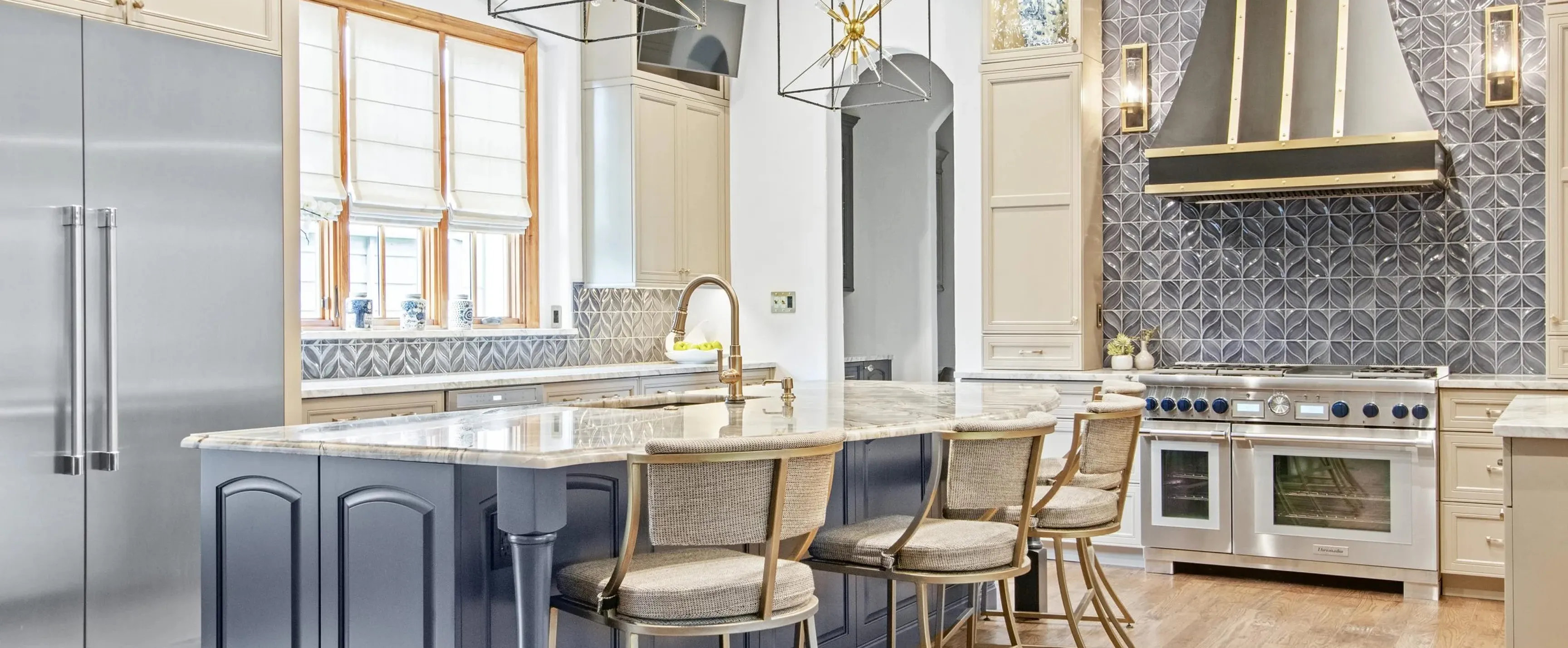 Modern kitchen with marble island, four cushioned stools, stainless steel fridge and stove, decorative tile backsplash.