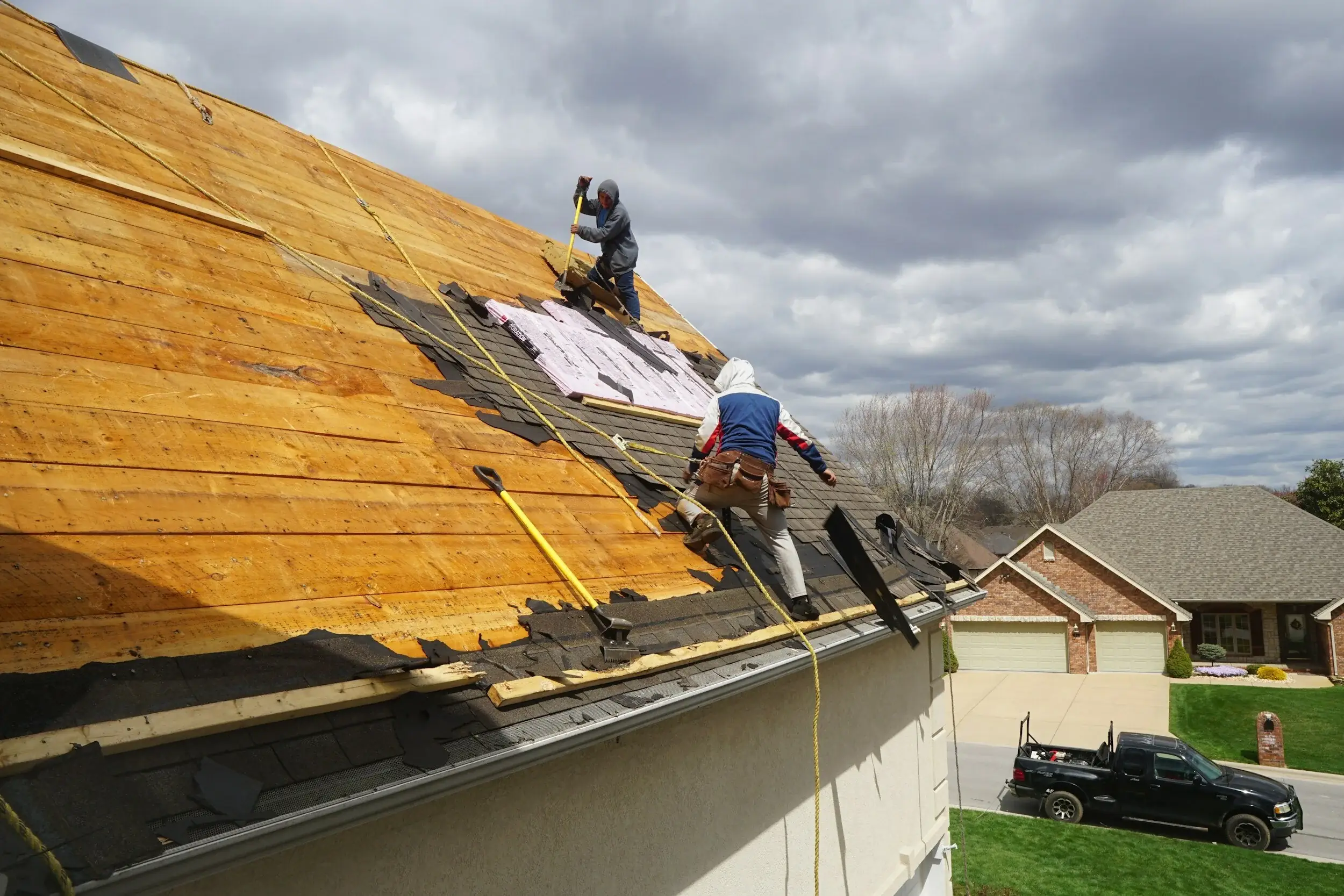 Two workers installing roofing shingles on a steep roof under a cloudy sky.
