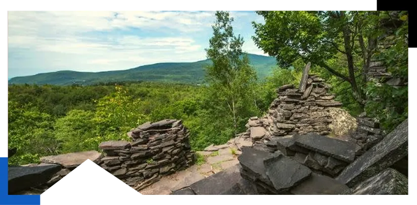 Stone ruins surrounded by green trees with hills and cloudy sky in the background.