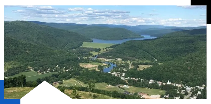 A valley with a winding river, surrounded by green hills and a small town under a cloudy blue sky.
