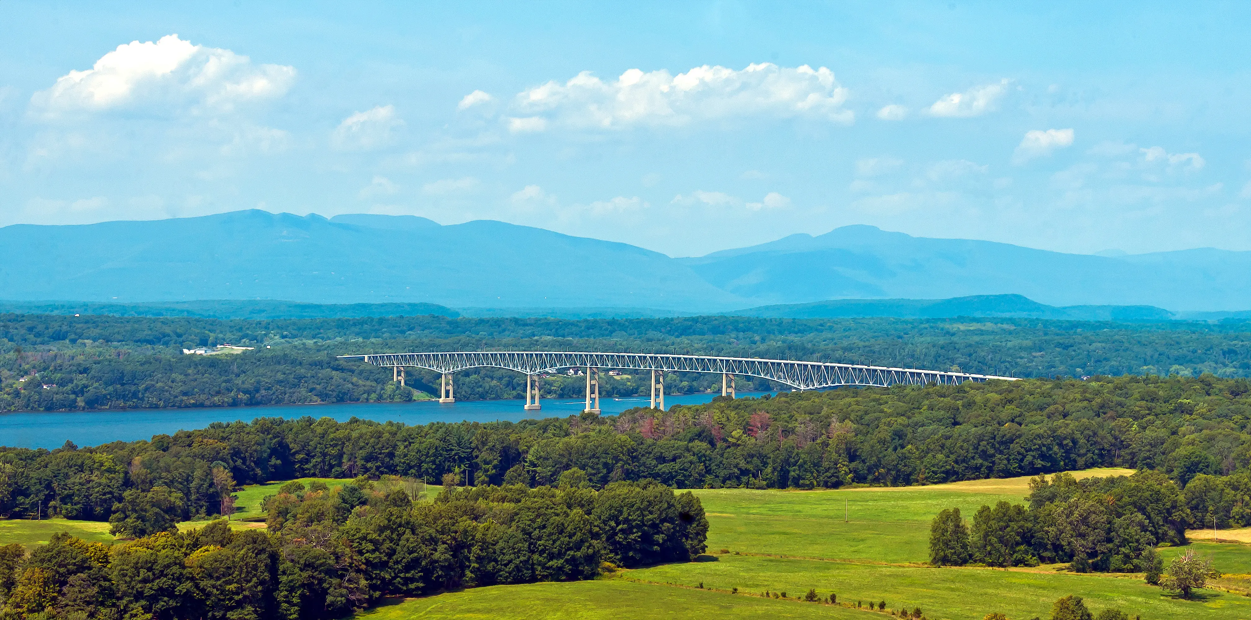 Long steel bridge crossing a river with forested land and mountains in the background under a blue sky.