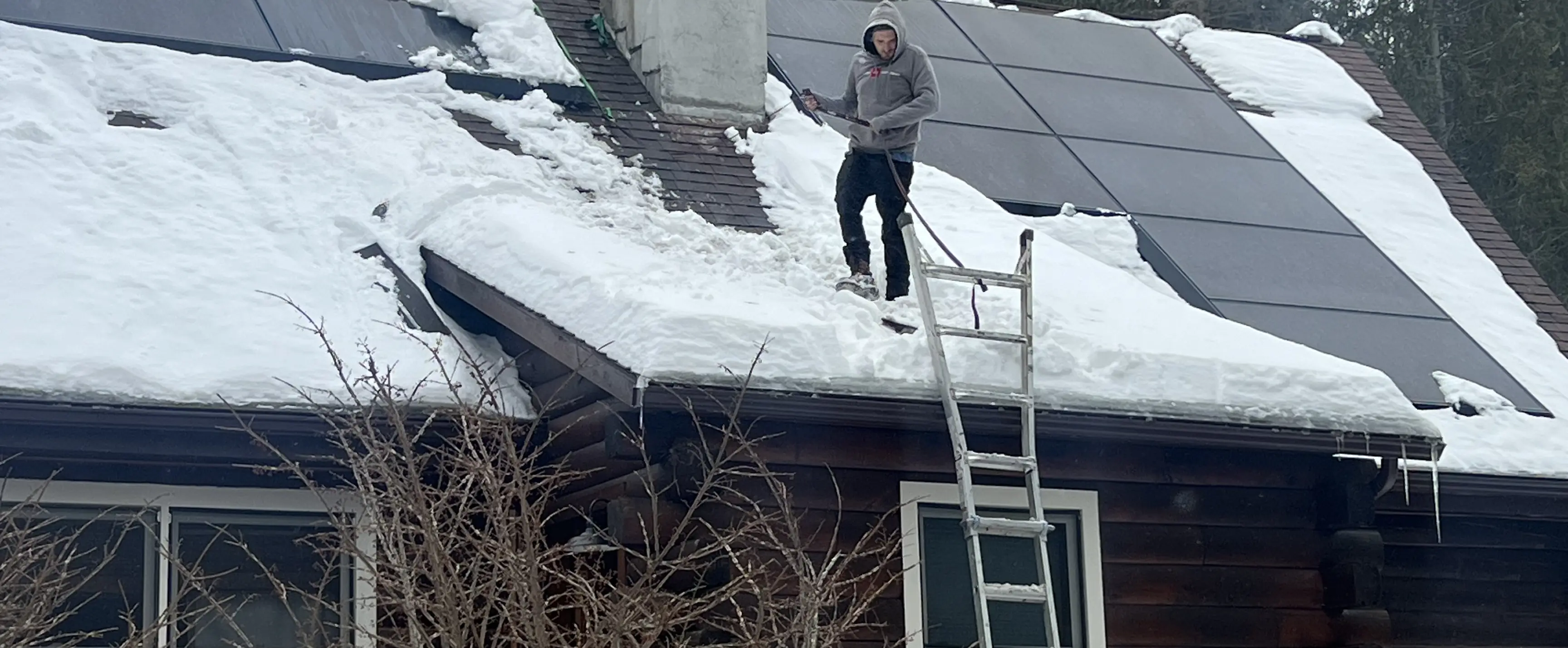 Man in hoodie clearing snow from roof with solar panels using a shovel and ladder.