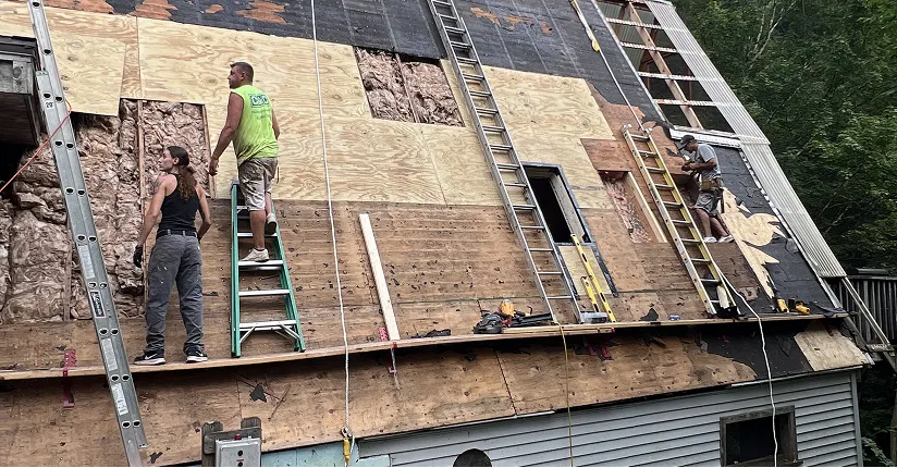 Three workers installing plywood on a steep roof with ladders and tools in a wooded area.