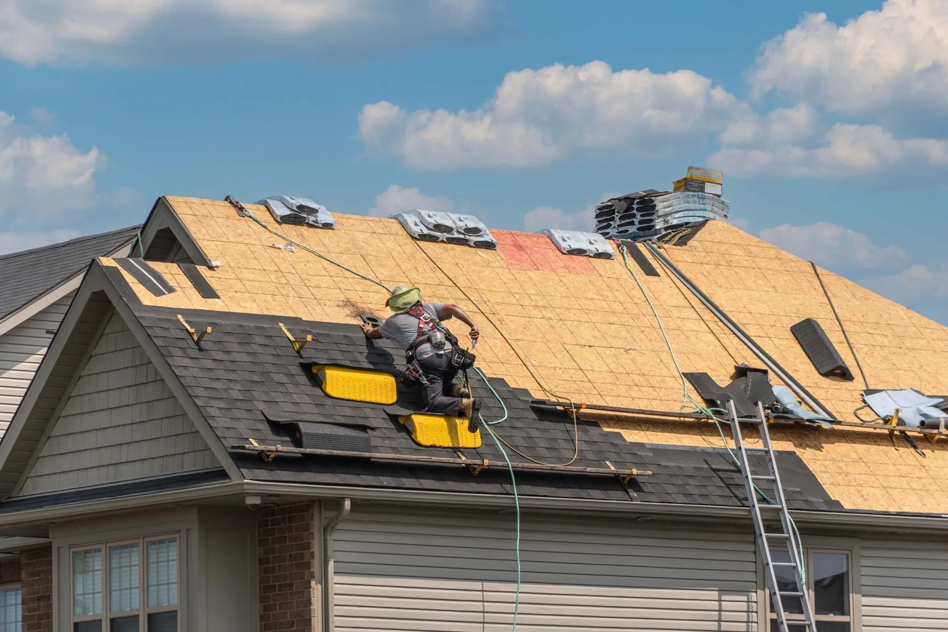 Worker installing new roof shingles on a house under a blue sky with scattered clouds.