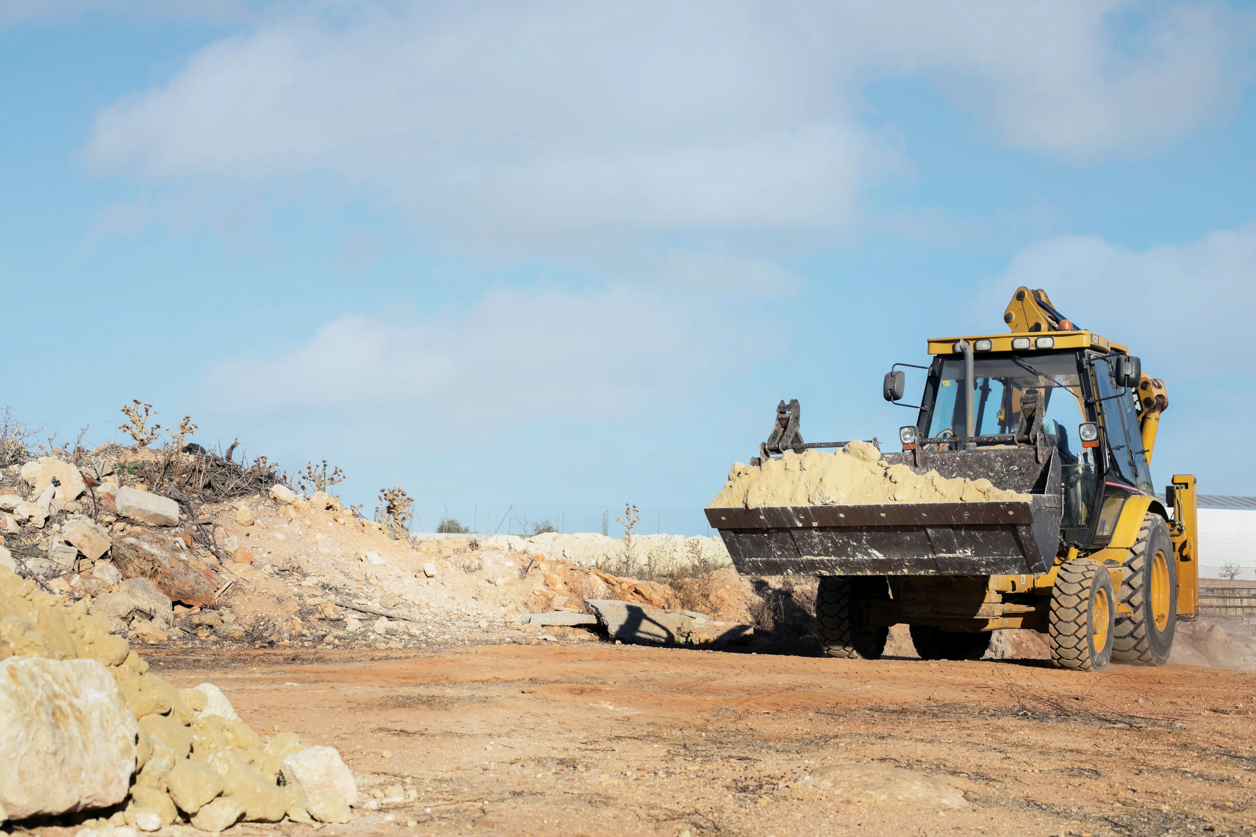 Yellow excavator with soil bucket driving on dusty construction site under blue sky.