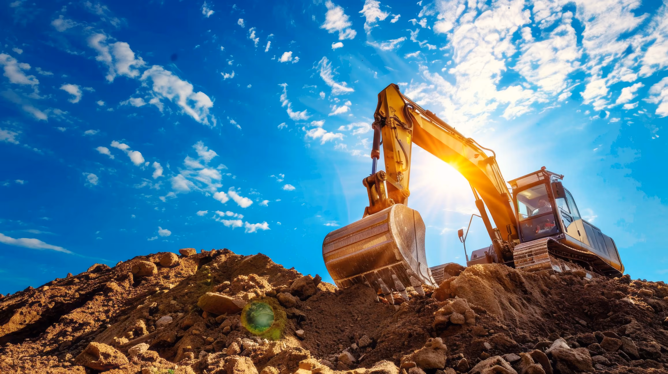 Large yellow excavator digging into a pile of dirt under a blue sky with scattered clouds.