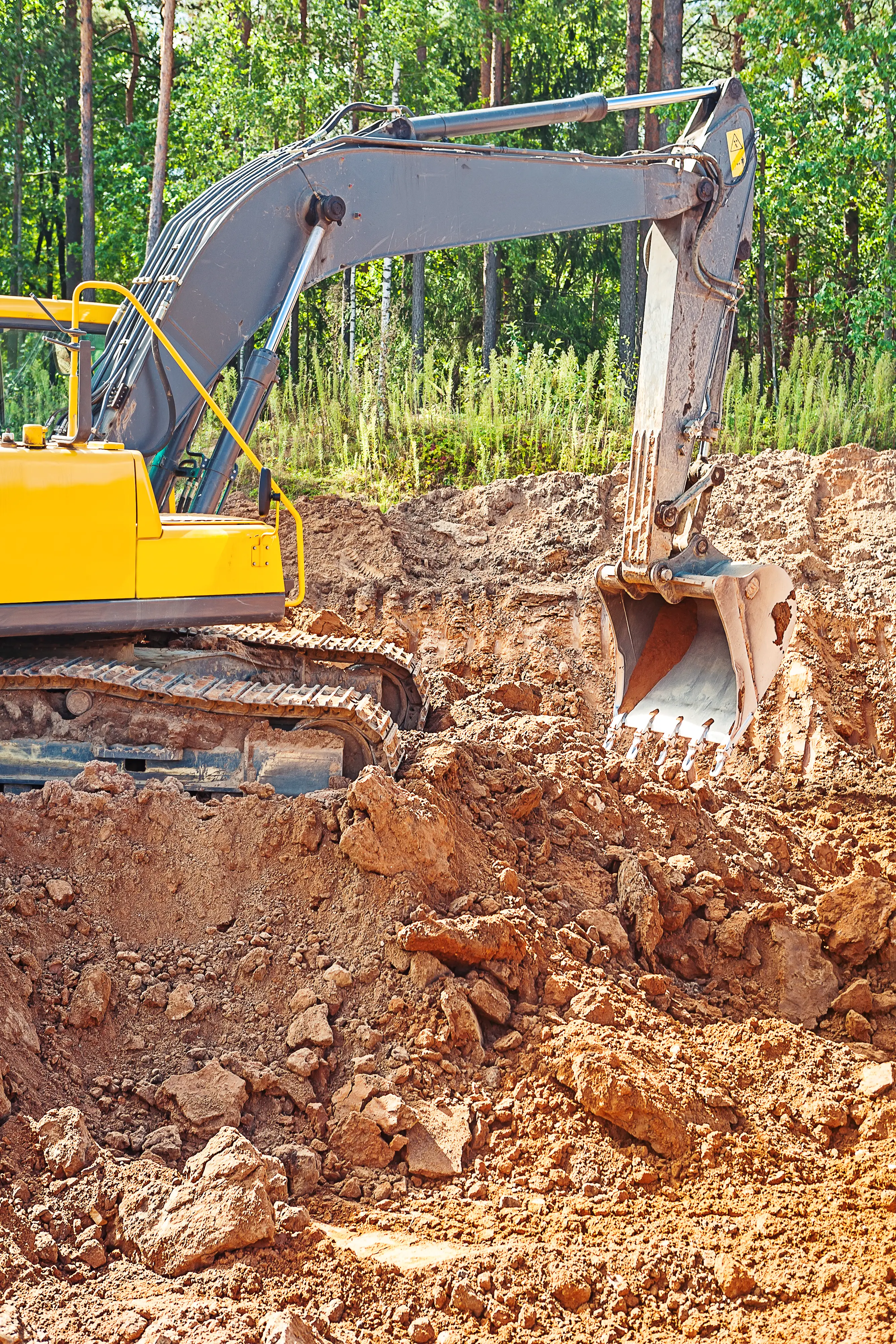 Yellow excavator digging clay soil at a construction site surrounded by trees in the background.