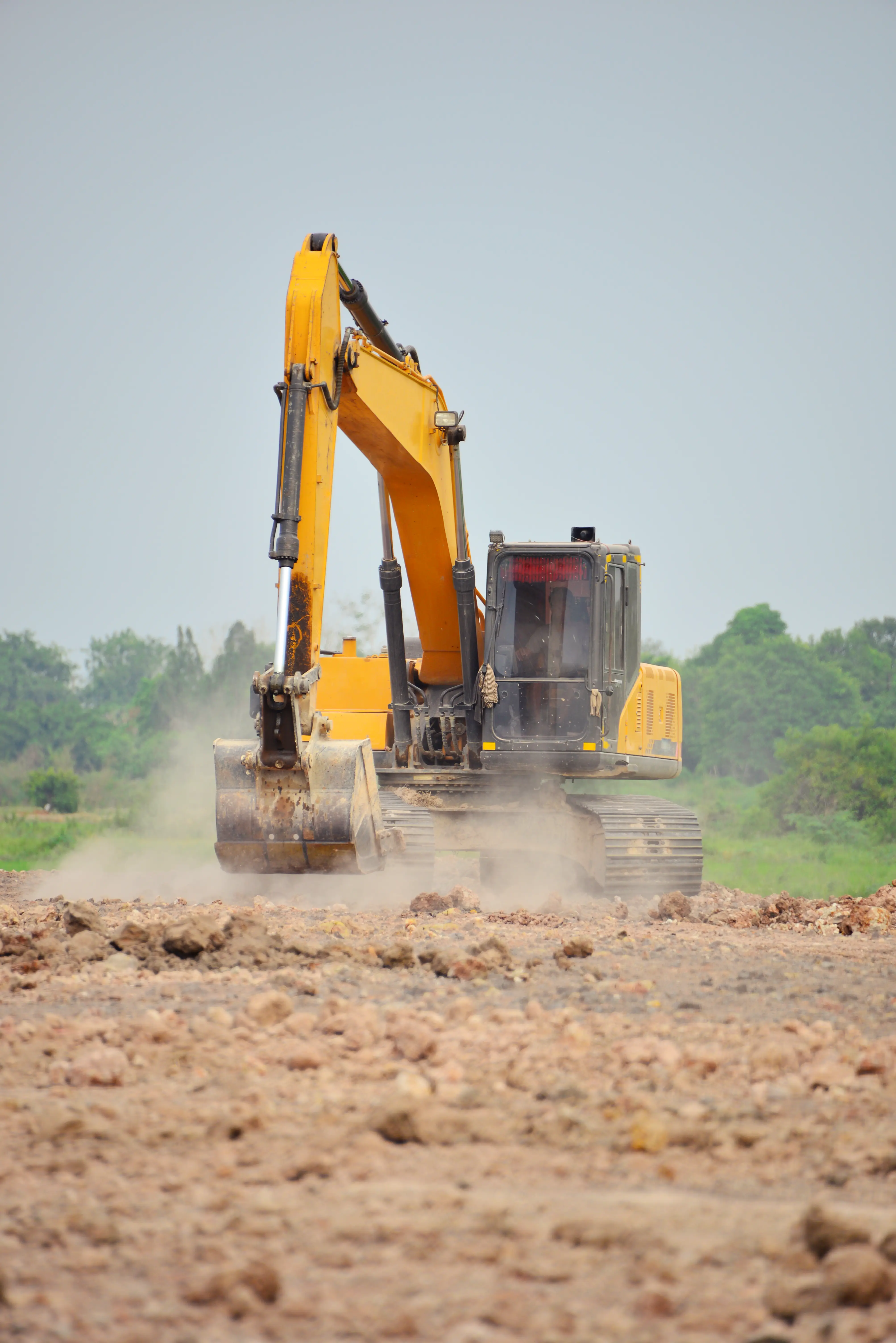 Yellow excavator working on a dusty construction site with trees in the background.