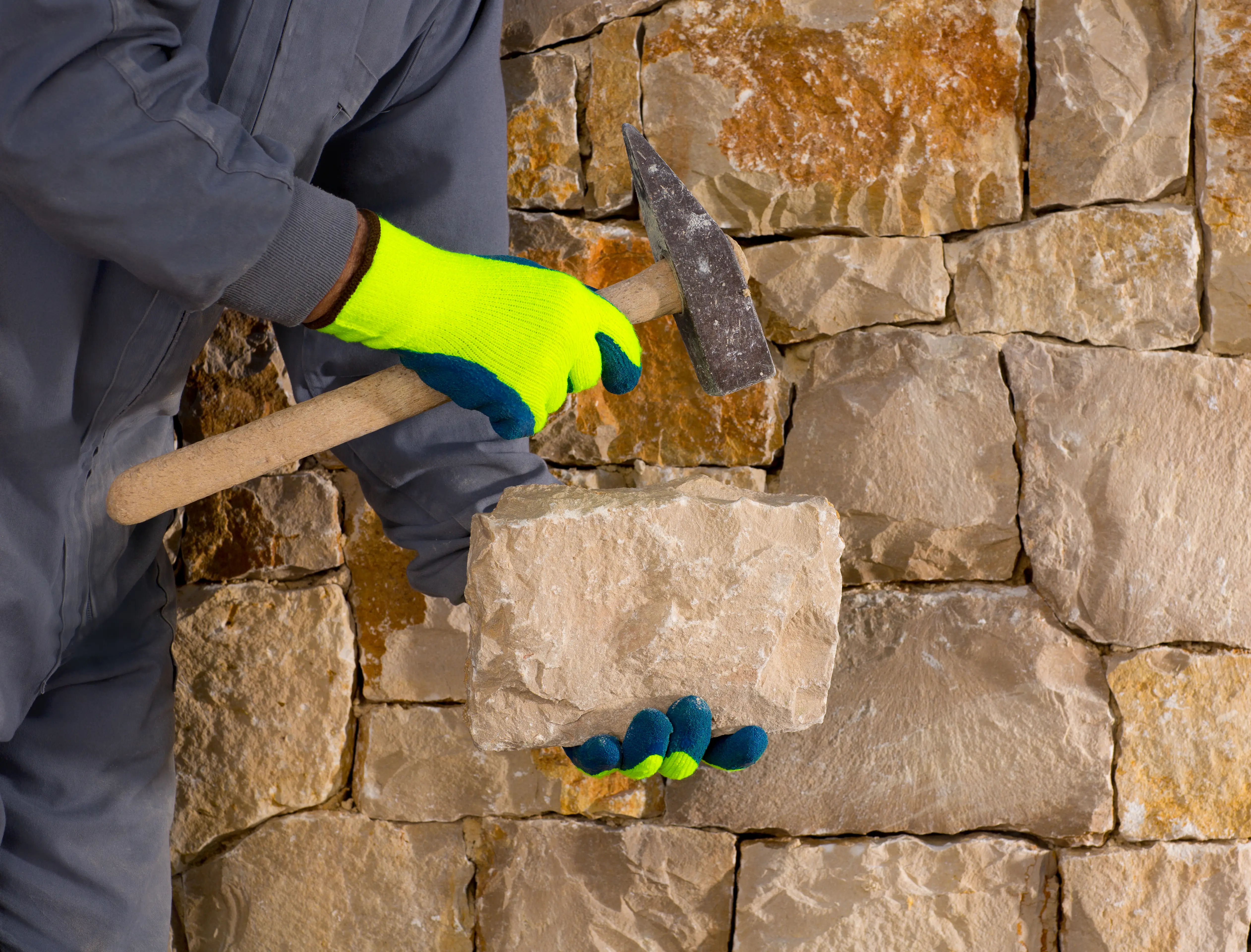Worker wearing gloves shaping a stone with a hammer in front of a stone wall.