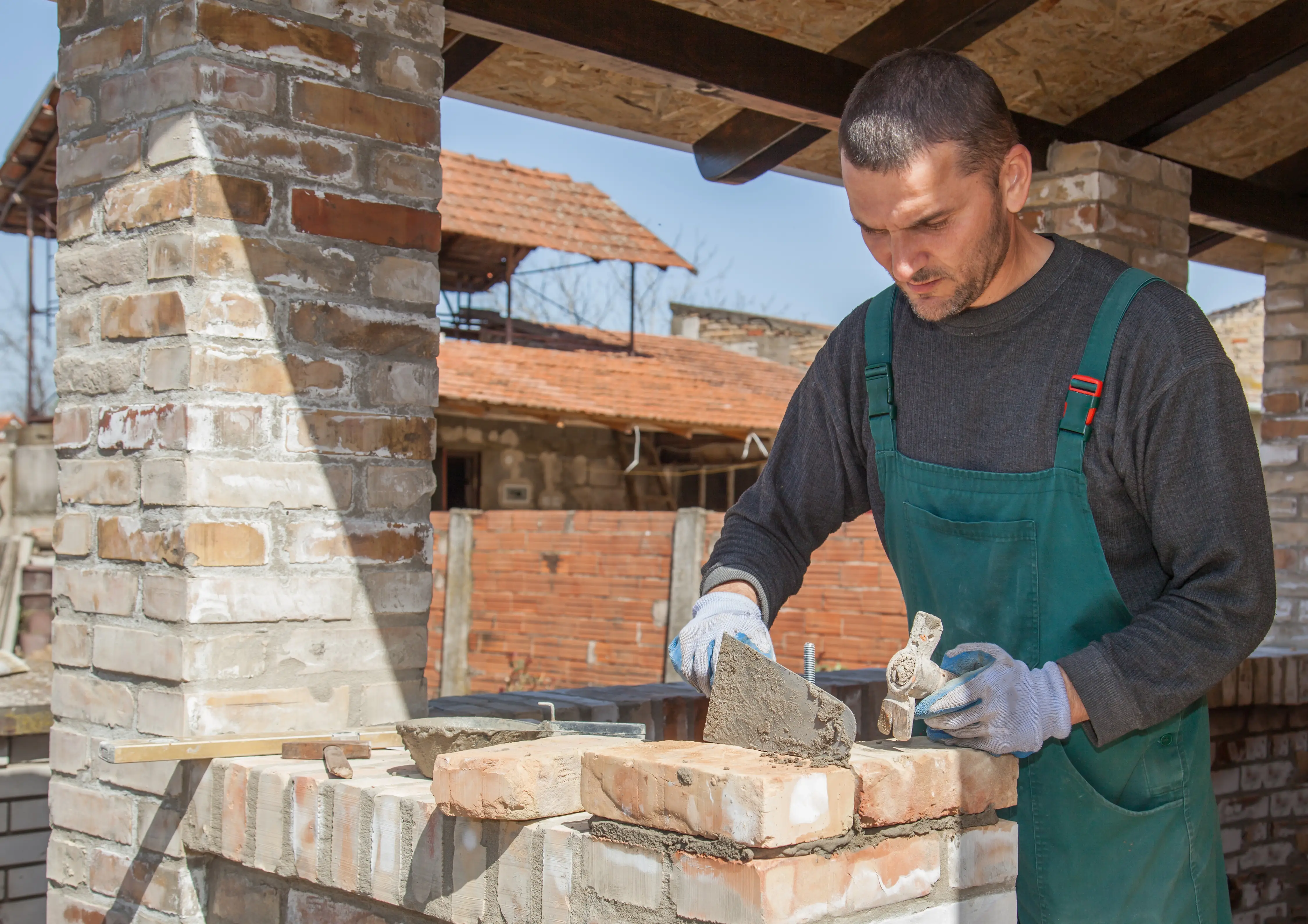 Man wearing green overalls applying mortar to bricks while building a brick wall outdoors.