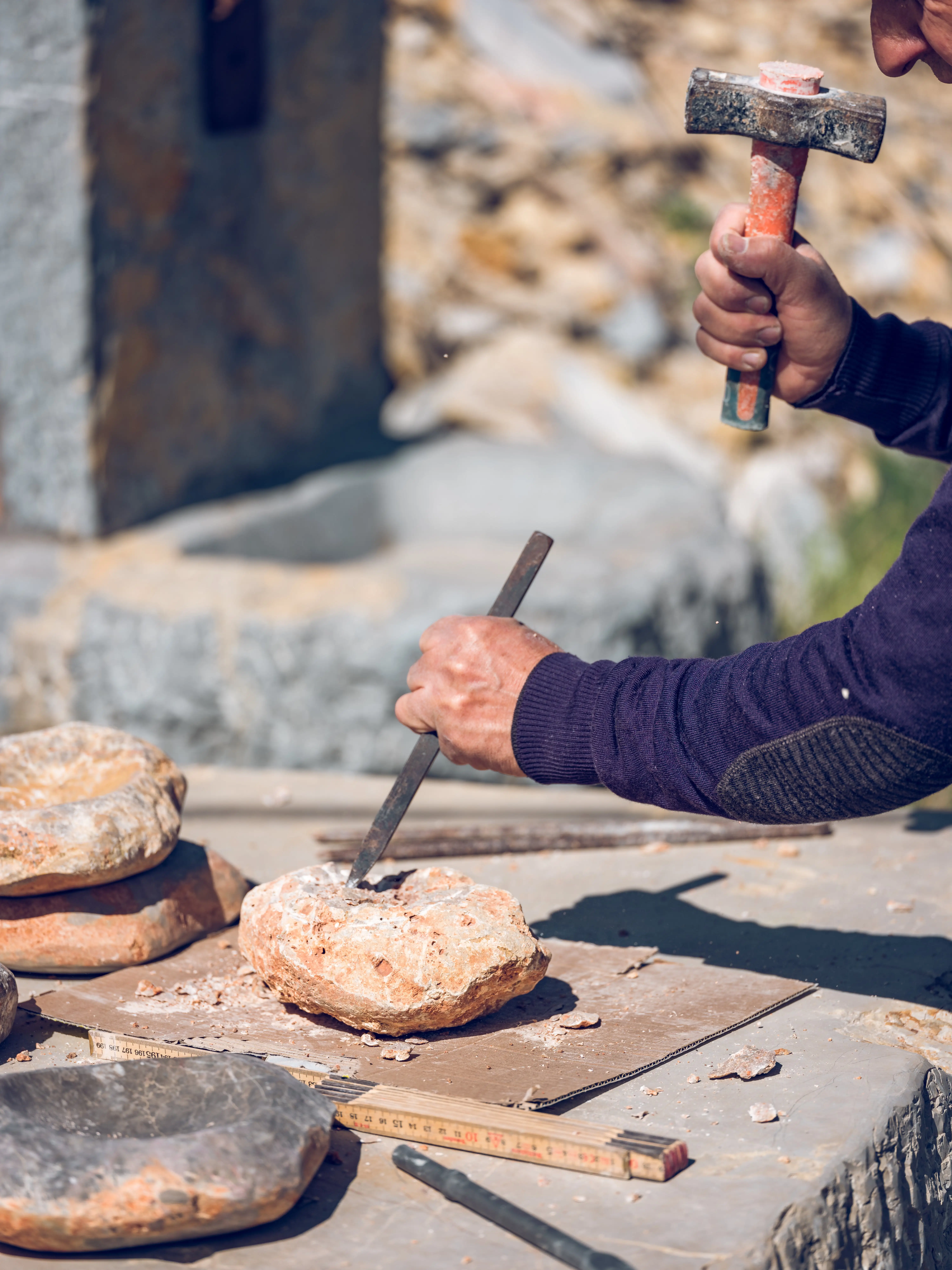 Person chiseling a large rock held on a table with other stones and tools around.