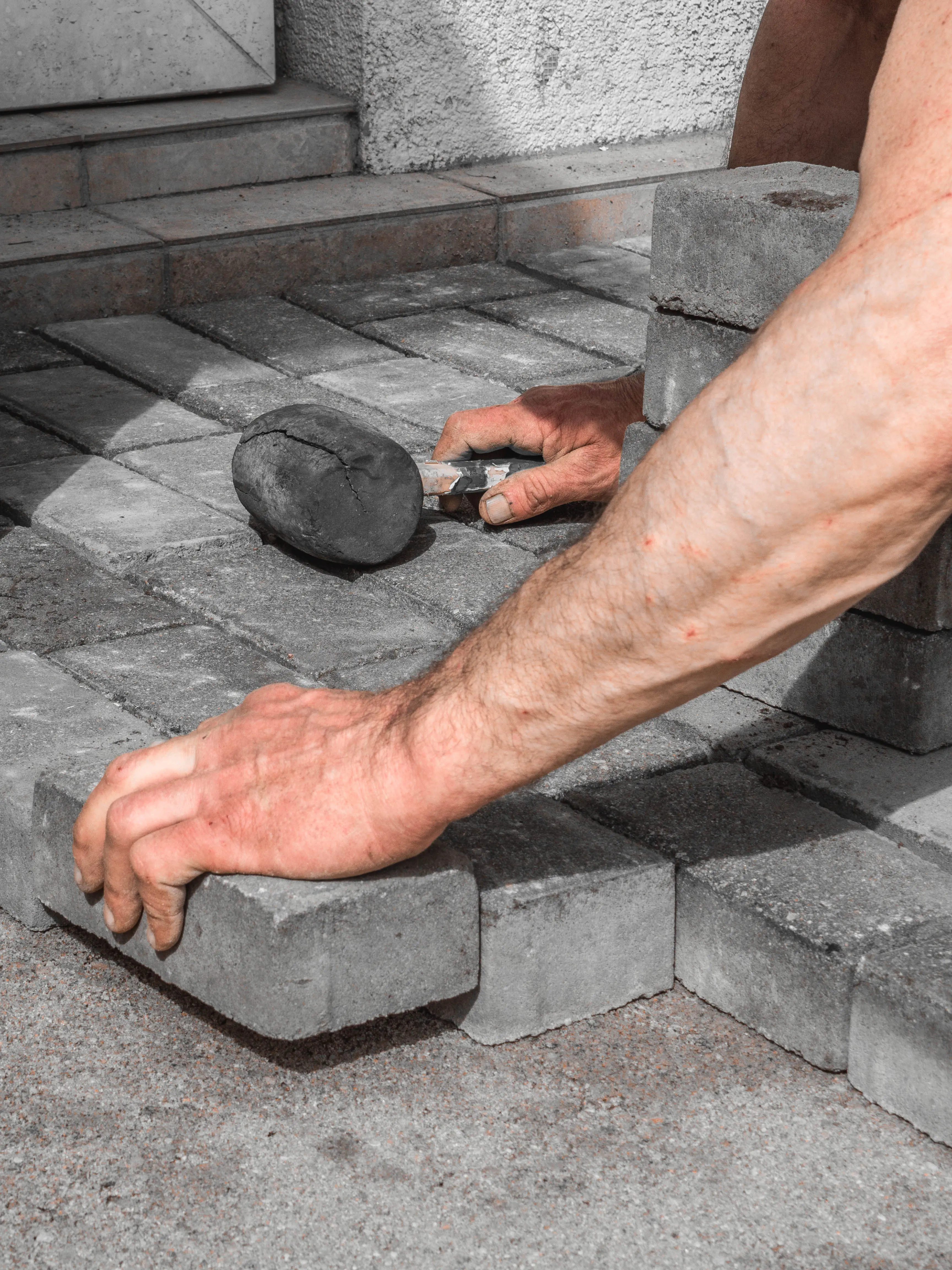 Hands installing square stone pavers on ground next to a rubber mallet and stacked stones.
