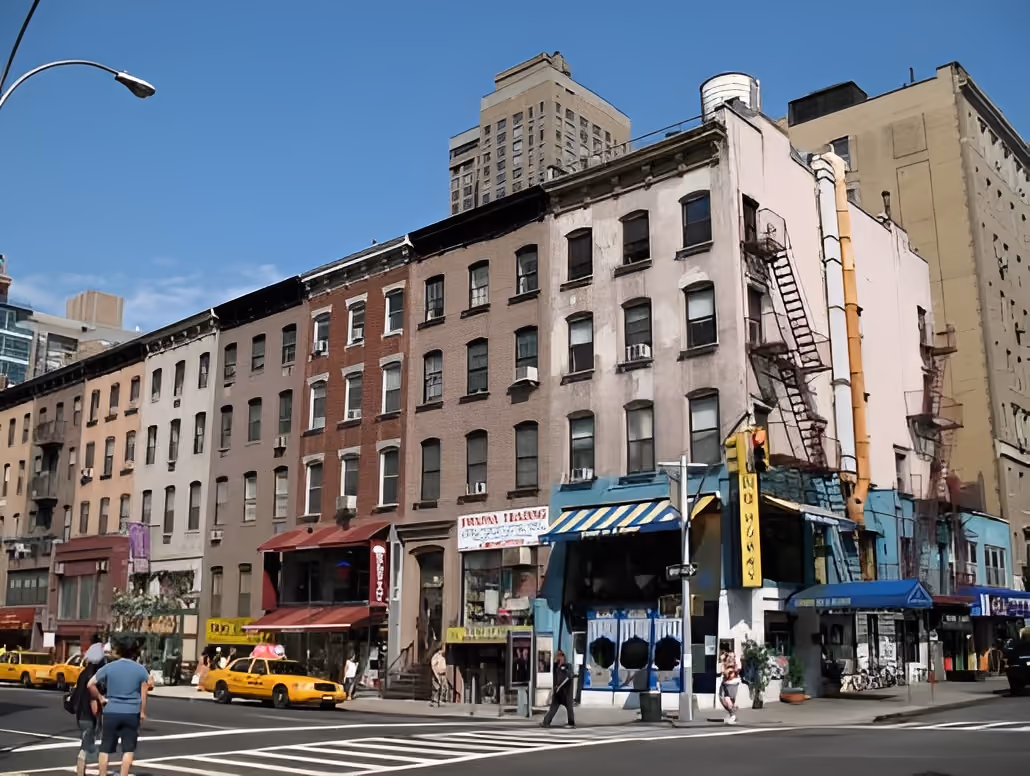 Street corner with multi-story brick buildings, shops, and yellow taxis under a clear blue sky.