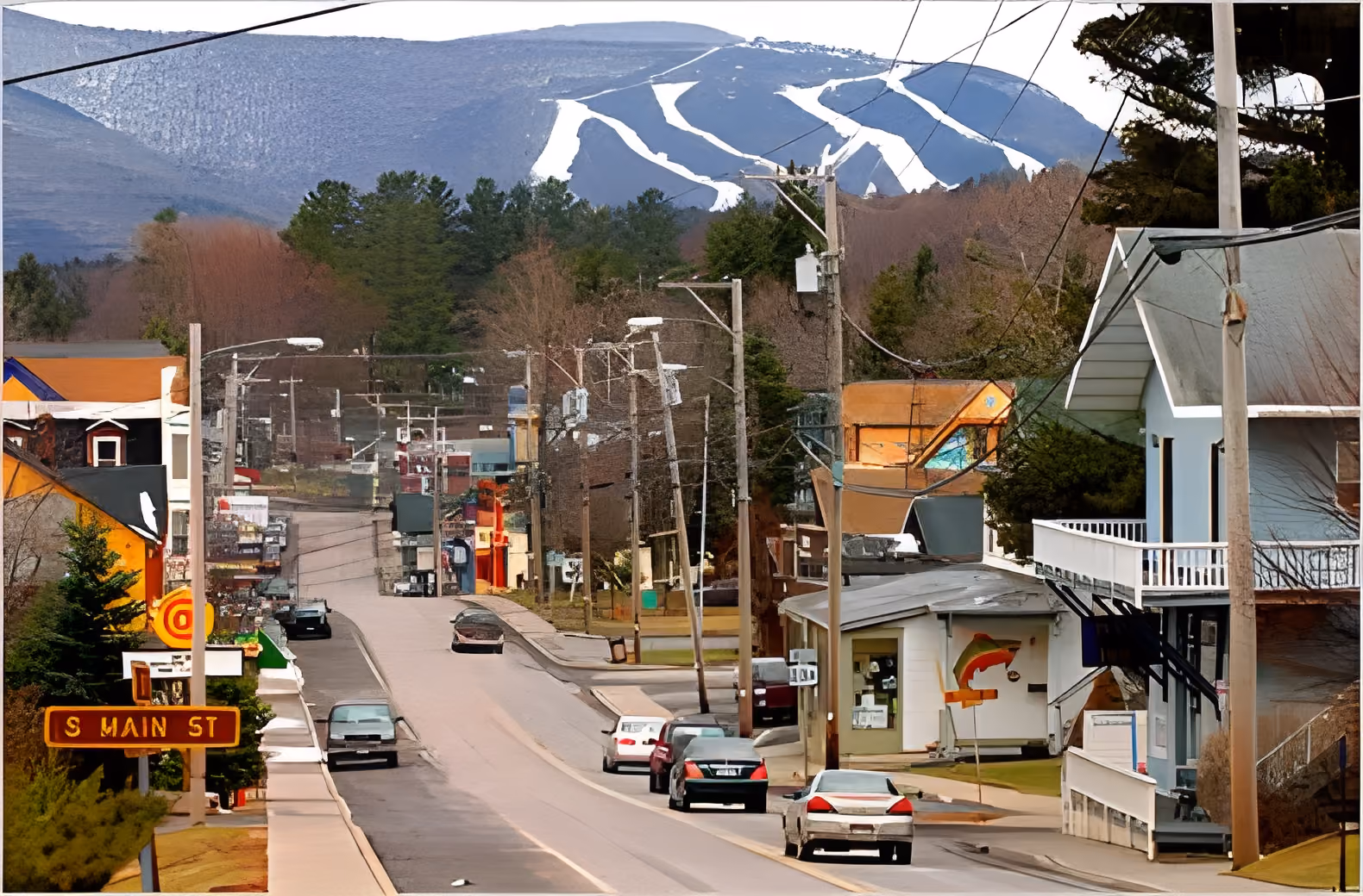 Small town street with cars and power lines, forested hills, and ski trails on mountain in background.