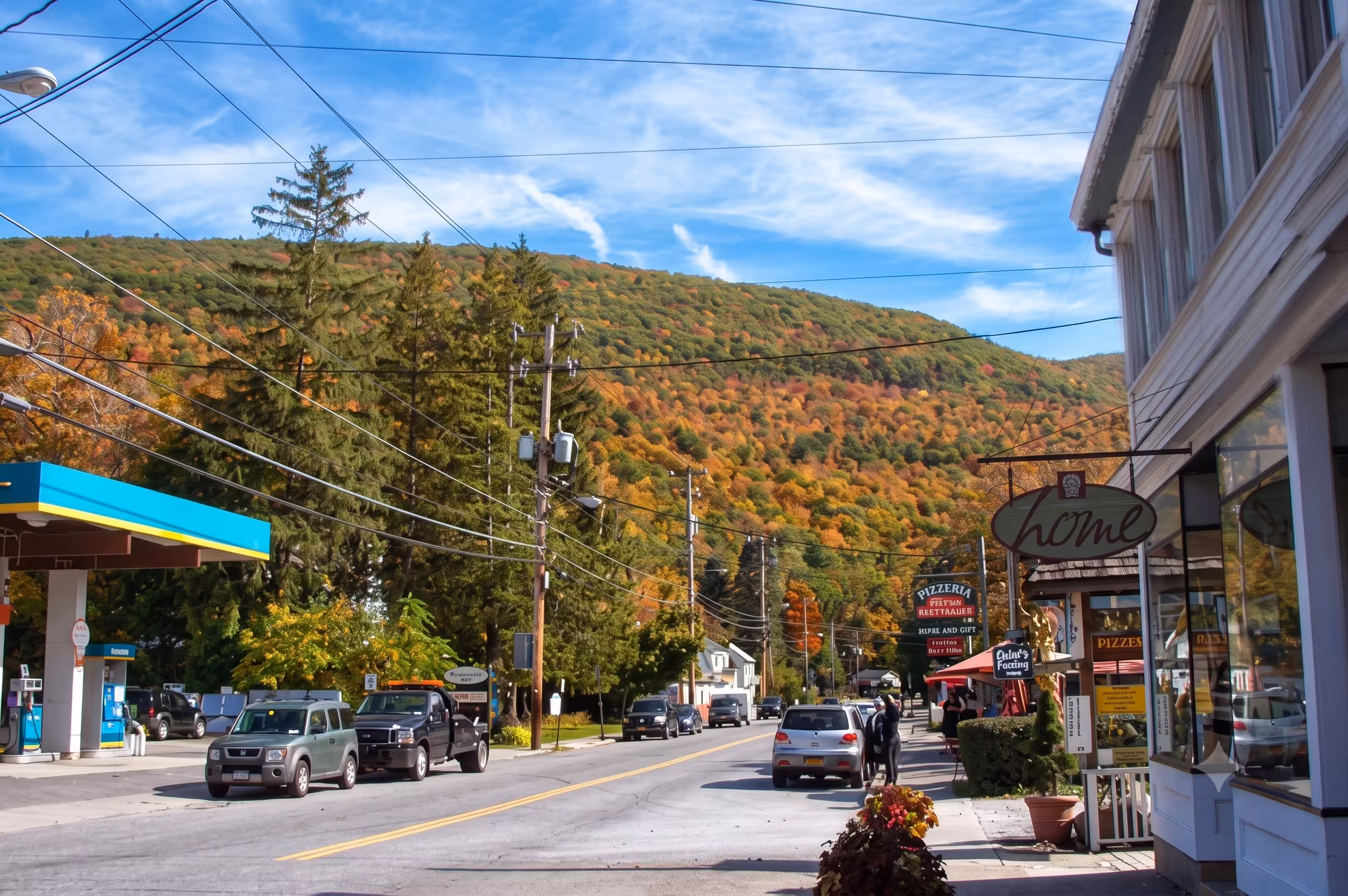 Small town street with cars, gas station, shops, and a forested hillside with autumn foliage.